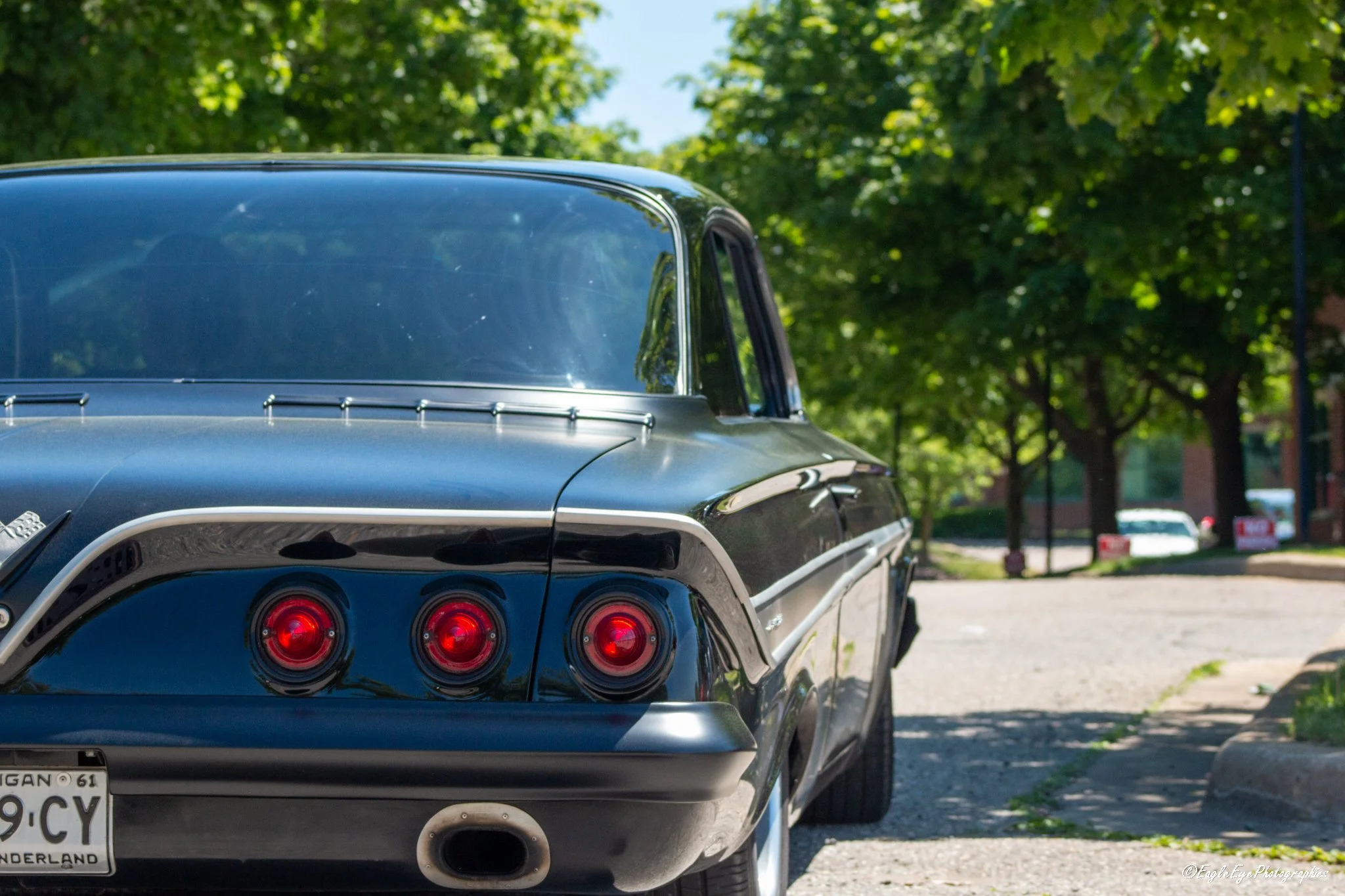 Rear view of a vintage black car with round red taillights parked on a street with trees and buildings in the background.