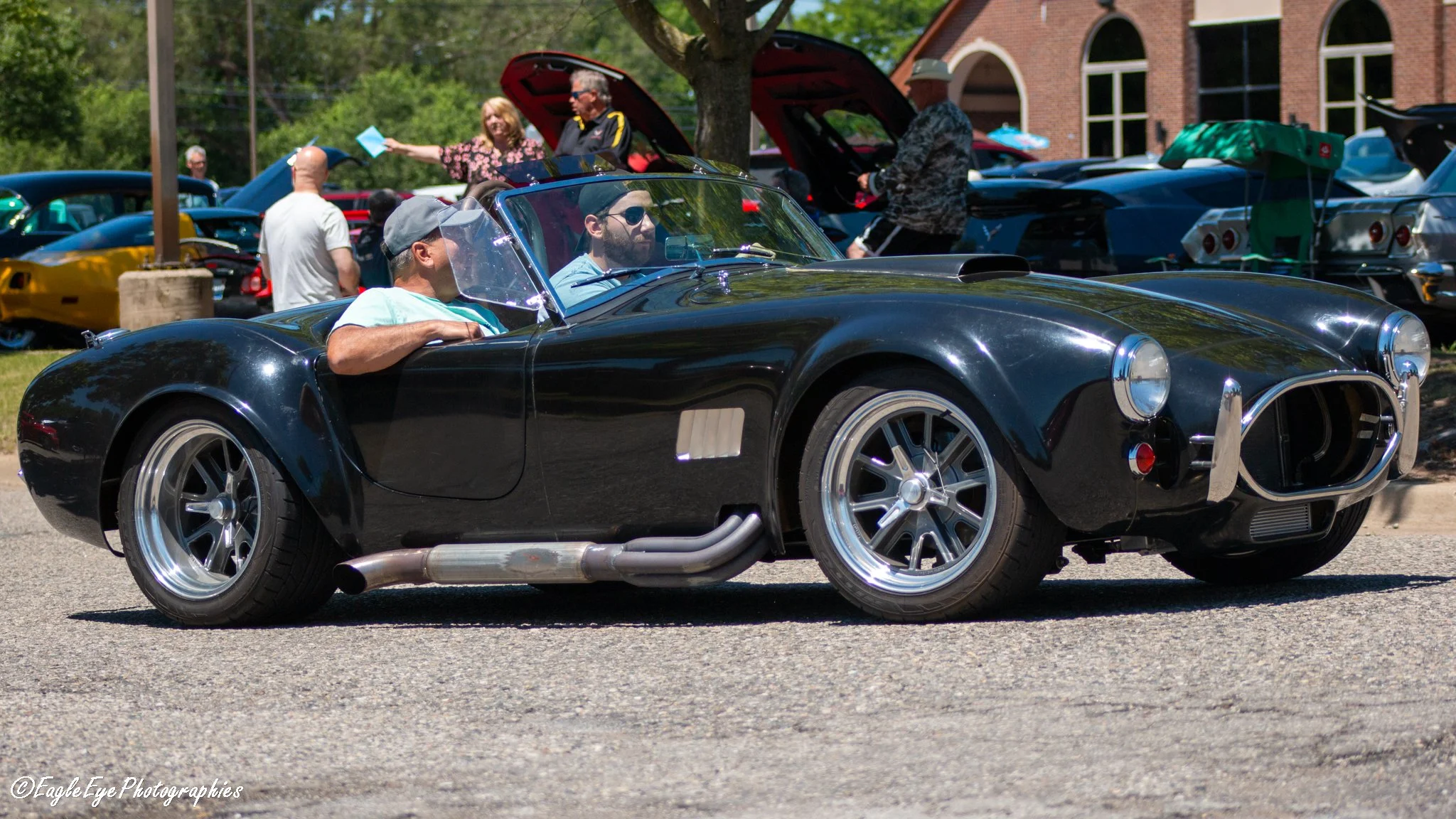 A black vintage convertible sports car parked at an outdoor car show, with people around it and open hood showing the engine, during daytime.