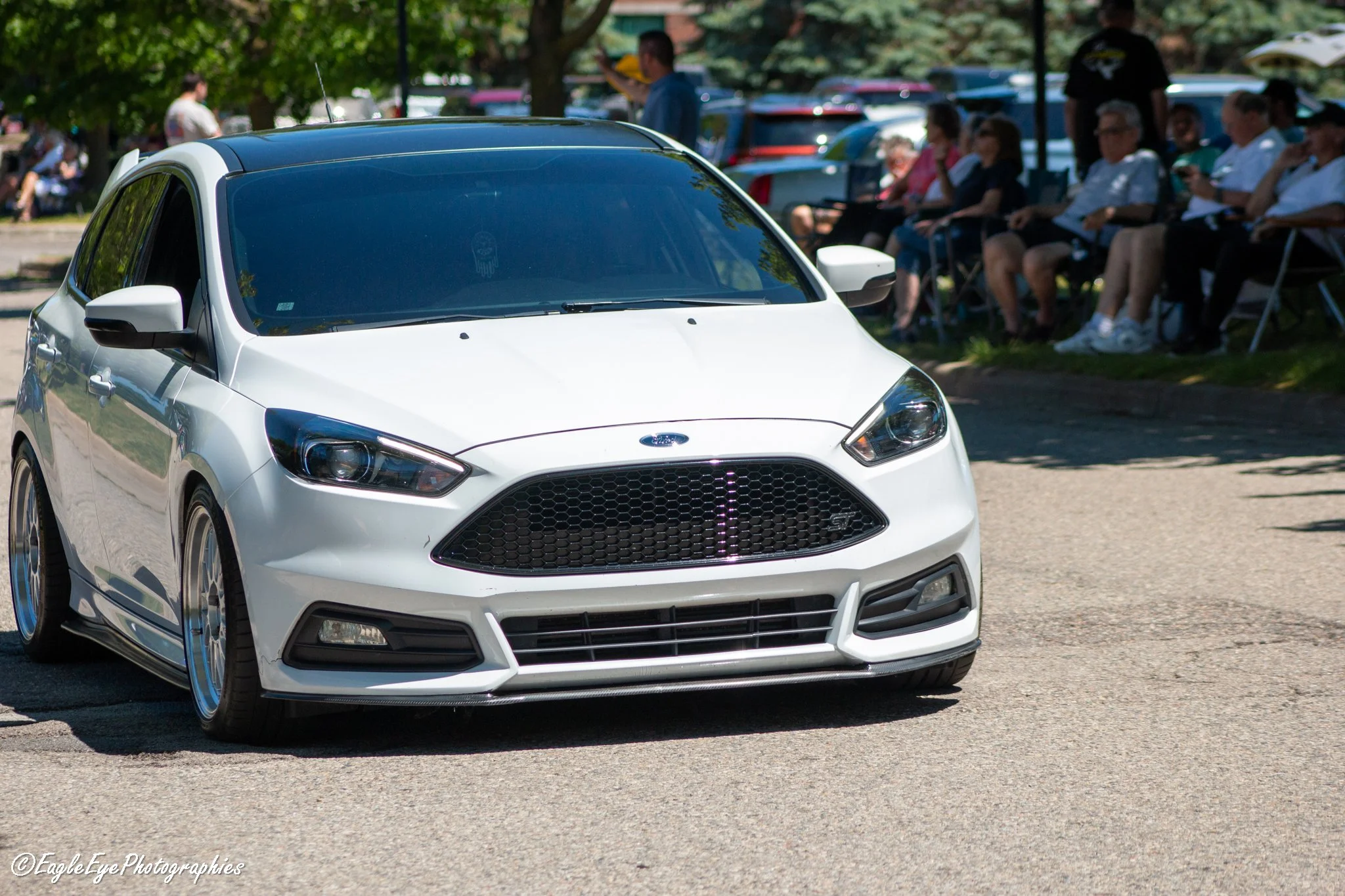 White Ford Focus hatchback car parked outdoors with a group of people sitting on chairs in the background, under trees on a sunny day.