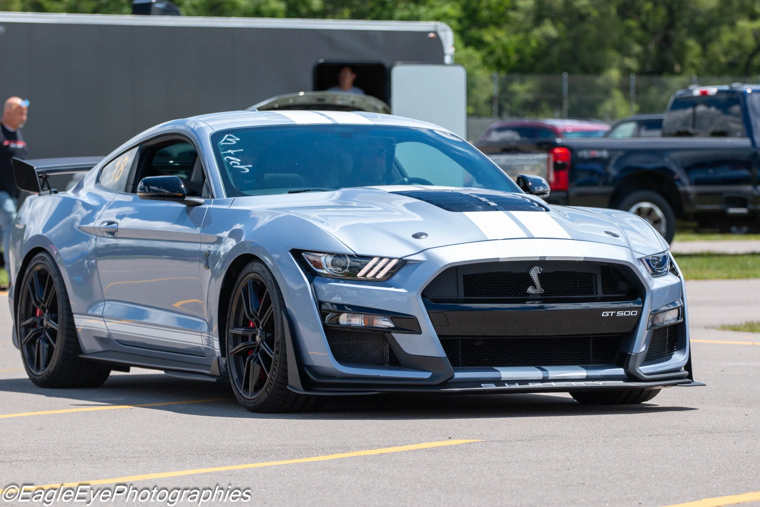 A silver Ford Mustang Shelby GT500 with black racing stripes parked in a parking lot, surrounded by other vehicles and people.