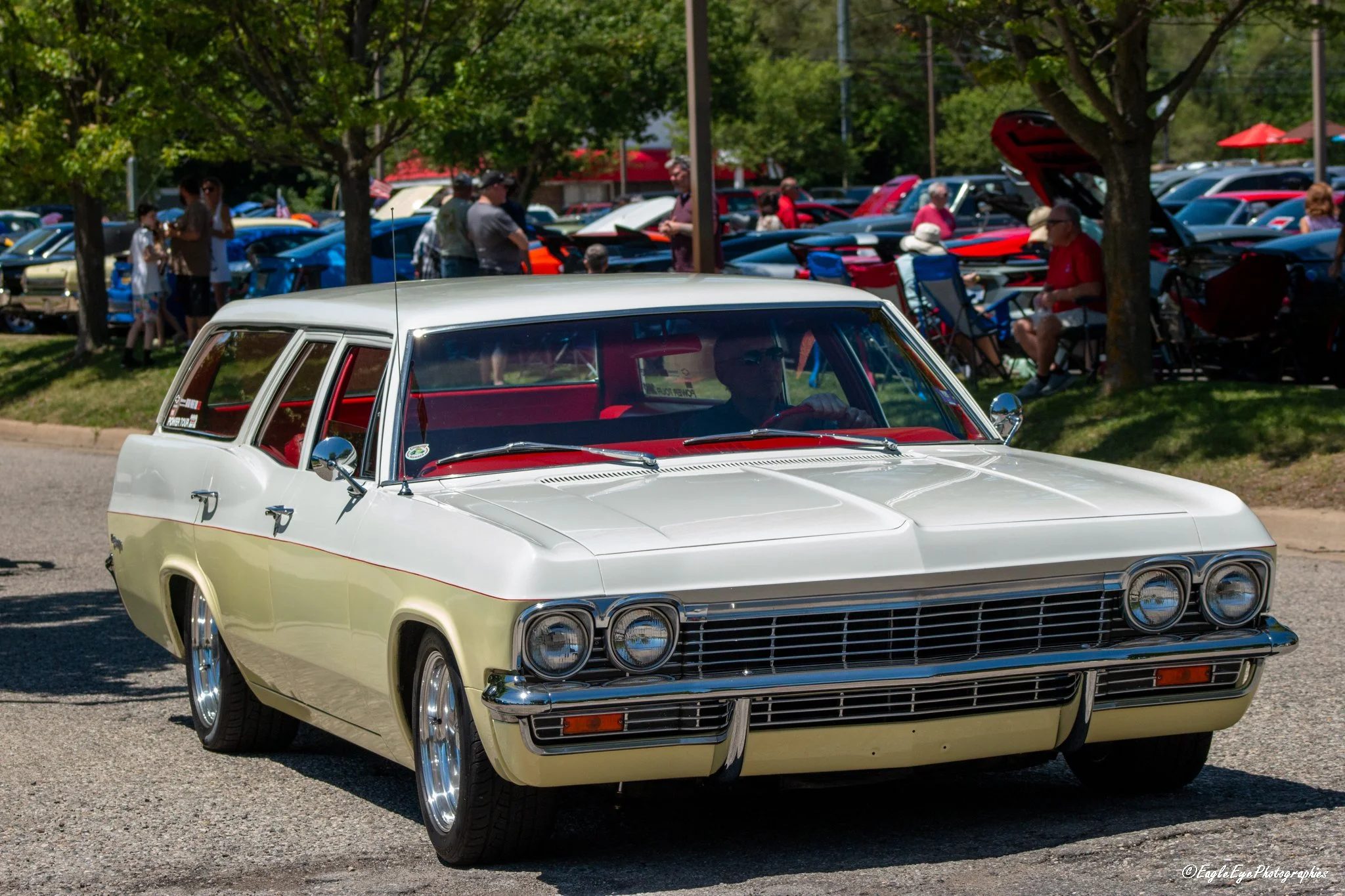 A vintage light-colored car parked in front of a crowd of people at an outdoor car show, with many classic cars and trees in the background.