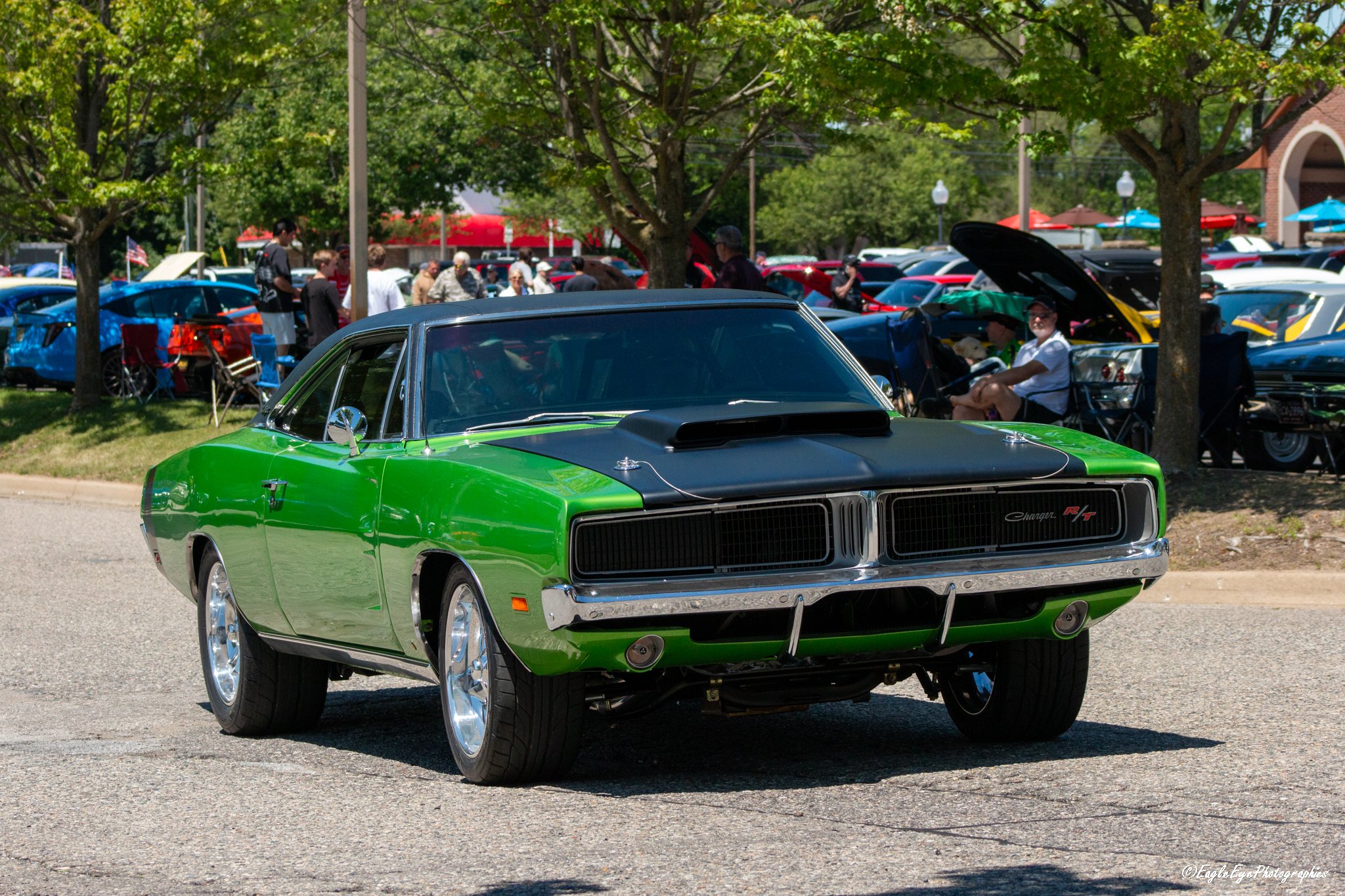 A bright green vintage muscle car with a black hood and chrome accents parked on a street during a car show. In the background, multiple tents, cars, and a crowd of people are visible under trees and a partly cloudy sky.