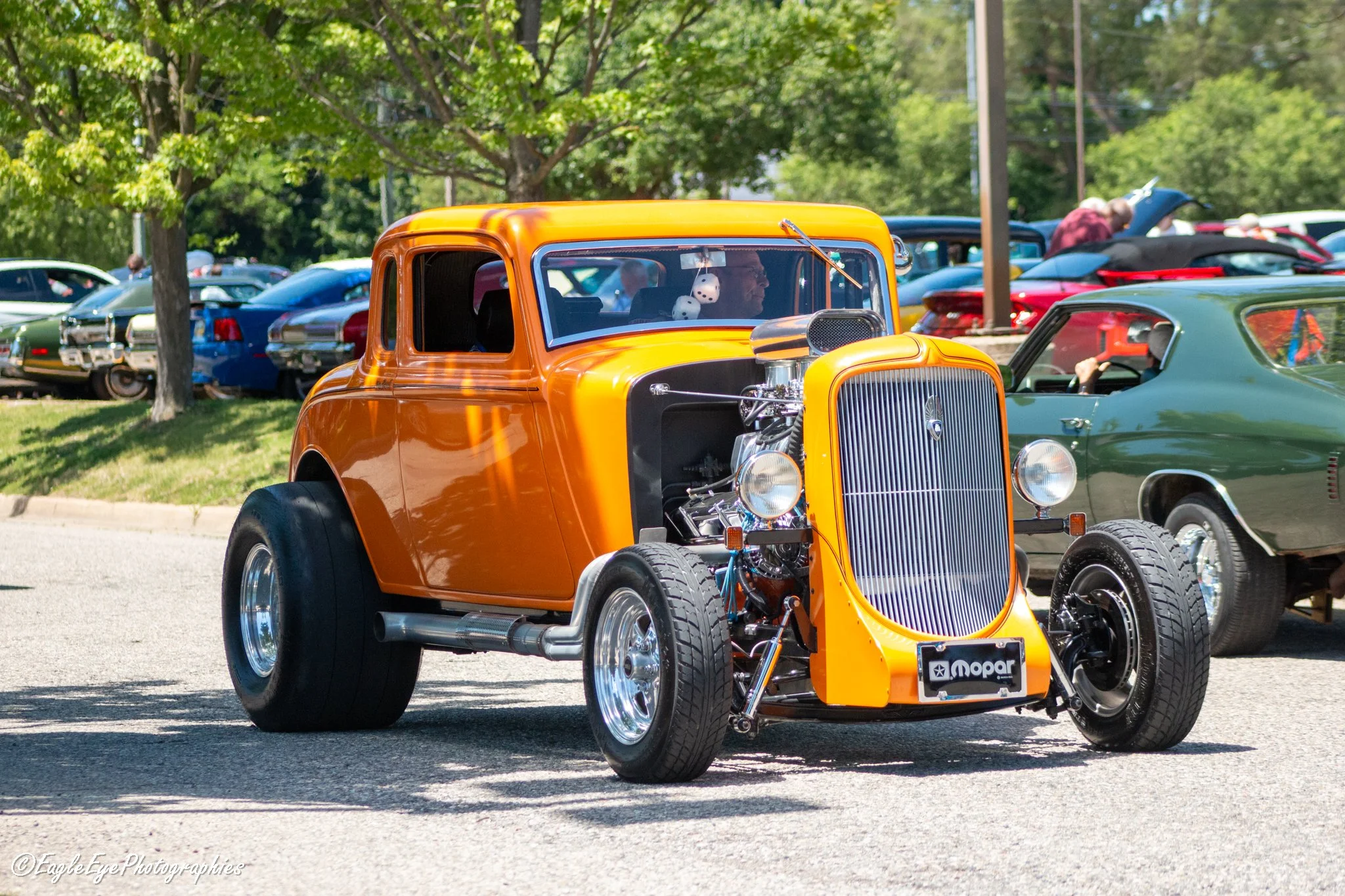 A vintage orange hot rod car with a visible engine, parked among other classic cars at an outdoor car show on a sunny day, trees in the background.