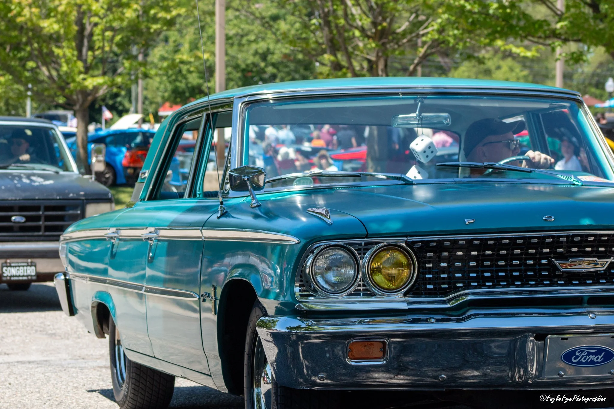 Close-up of a vintage teal Ford car with yellow-tinted headlights and chrome detailing, driving at a car show with other classic cars and people in the background.