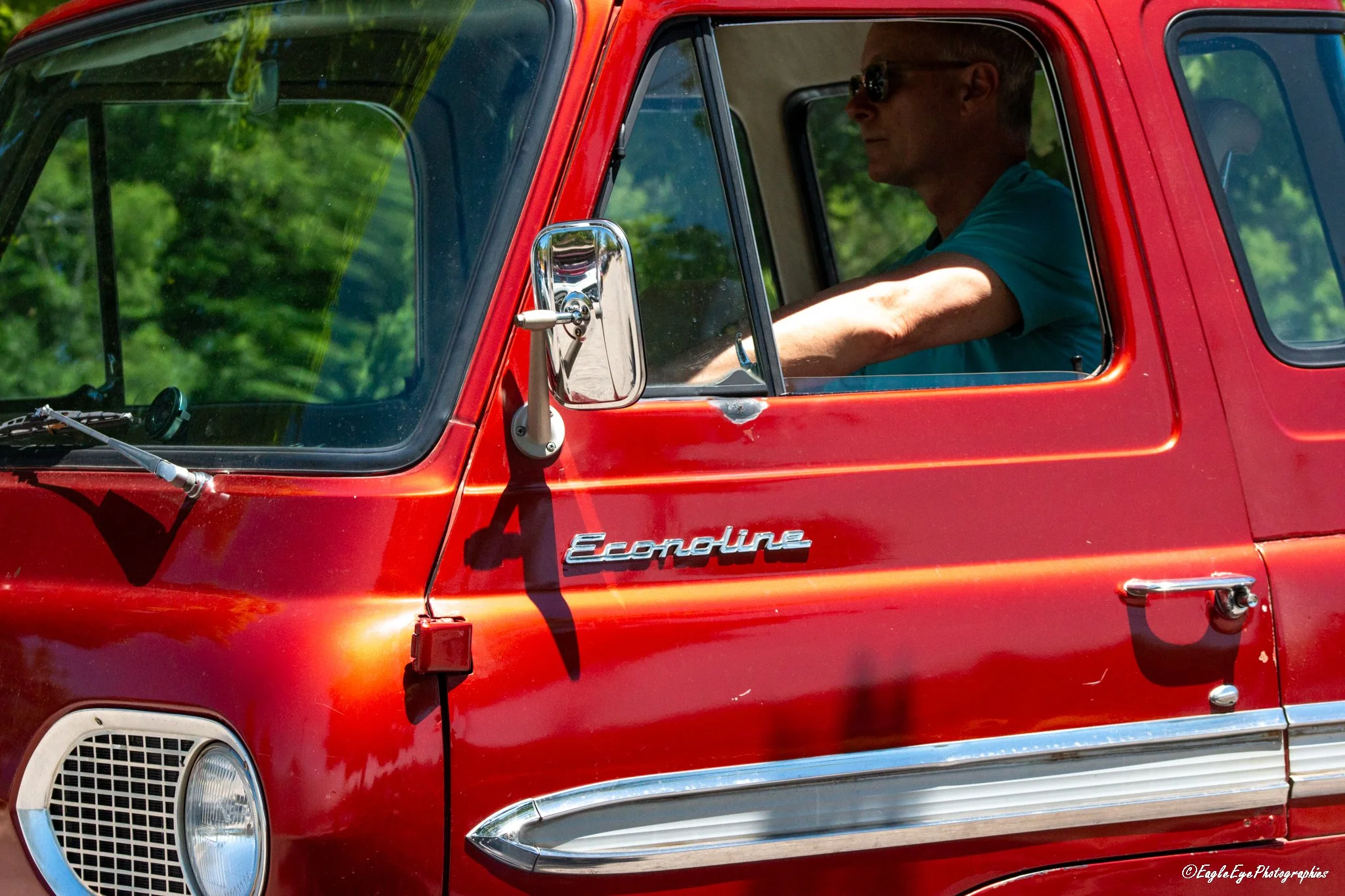 Close-up of the front side of a vintage red Ford Econoline van, showing the side mirror, part of the front grille, and the vehicle's logo, with a man wearing sunglasses sitting inside.