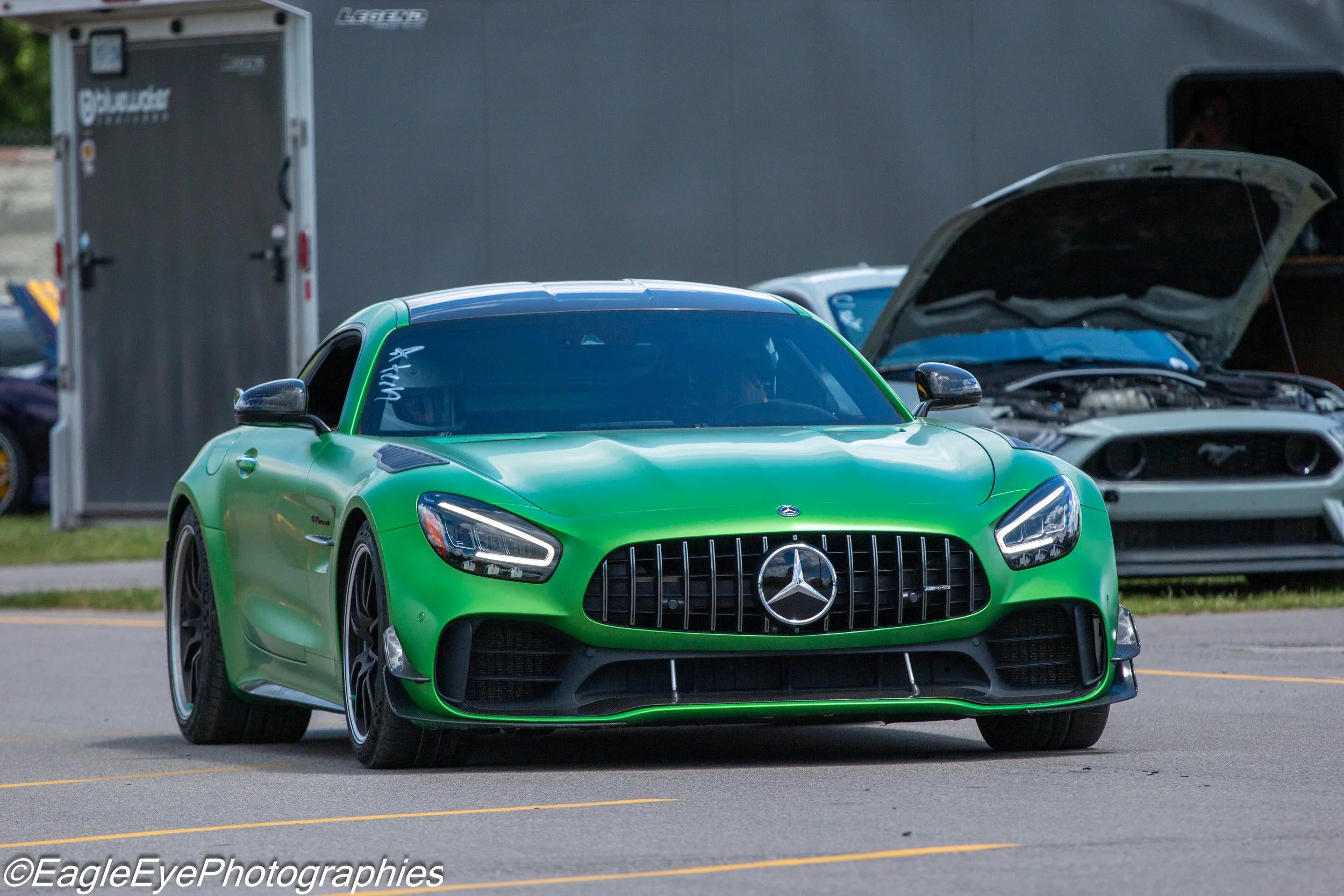A bright green Mercedes-Benz sports car with a black grille and rearview mirrors parked on a paved lot, with a trailer and other cars in the background.