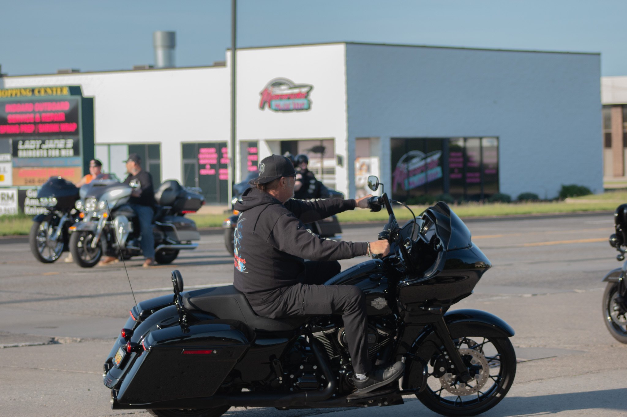 Multiple motorcyclists riding on a street in front of a white building with a pink and teal sign and a digital marquee board.