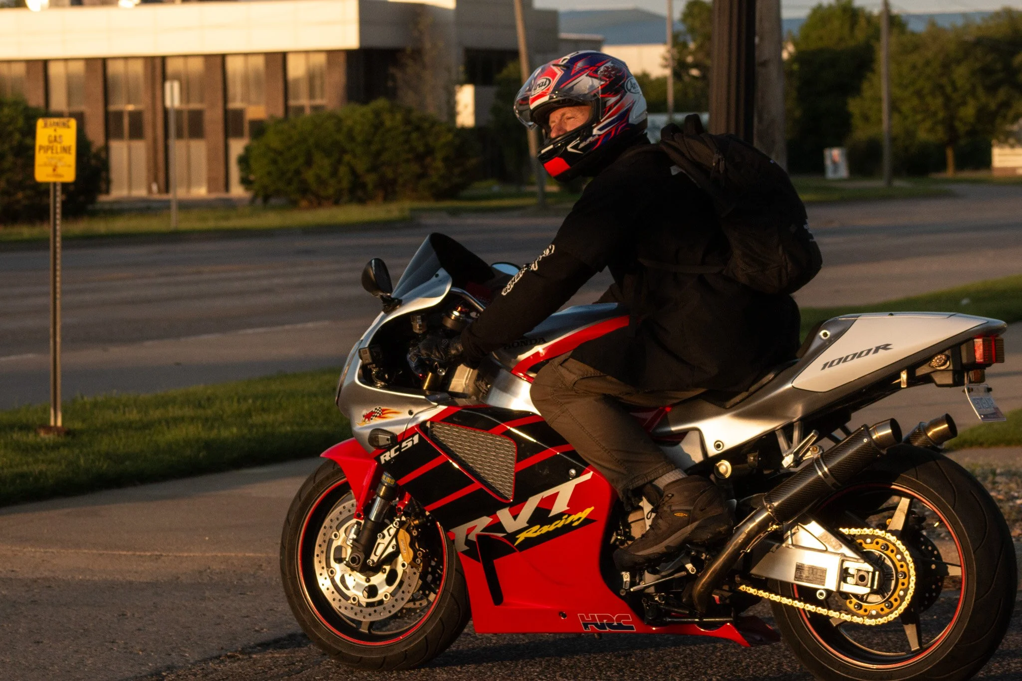 Person wearing a helmet and black riding gear sitting on a red and white sport motorcycle on a street in the evening.