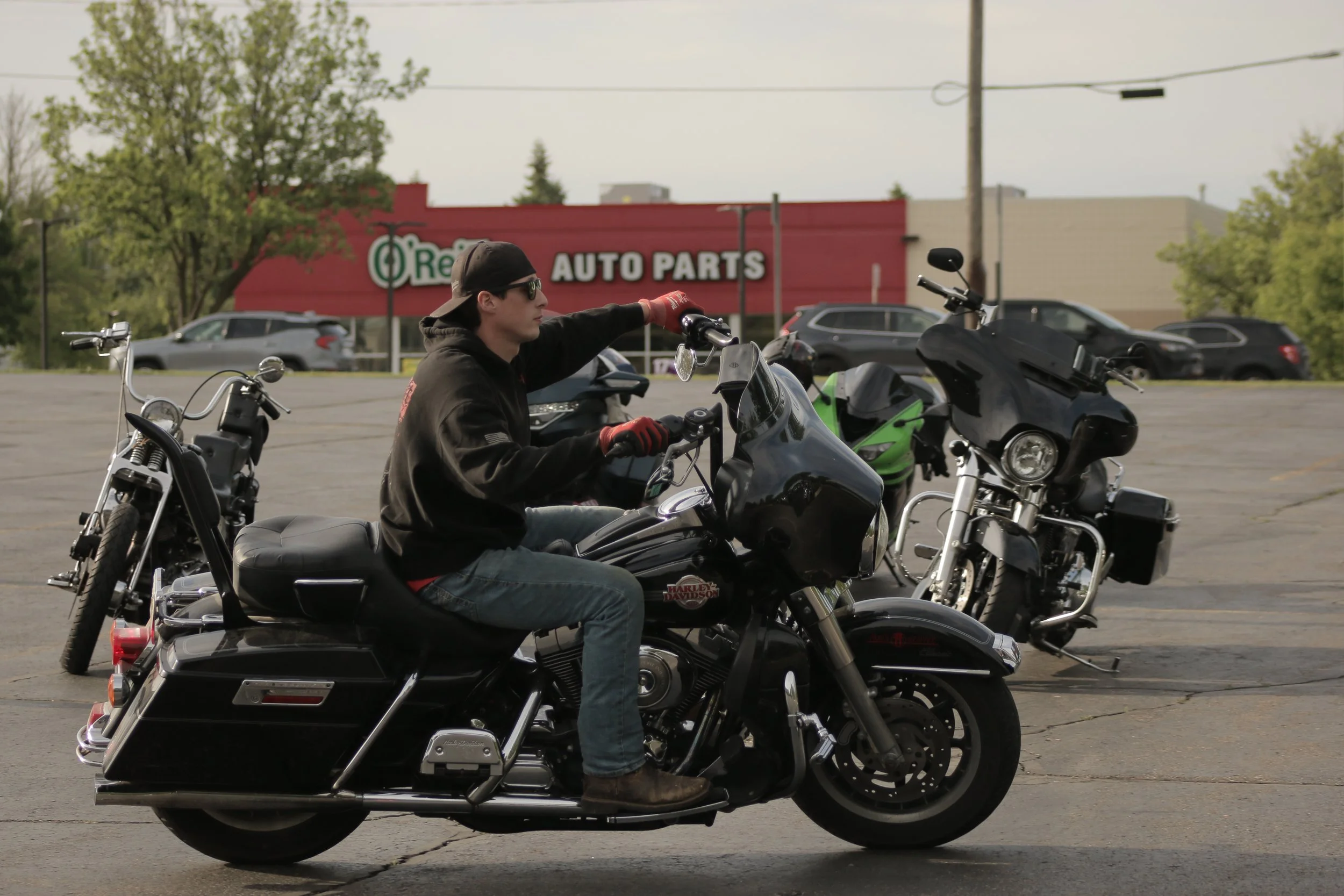 A man riding a black Harley Davidson motorcycle in a parking lot, with motorcycles parked around him and a red auto parts store in the background.