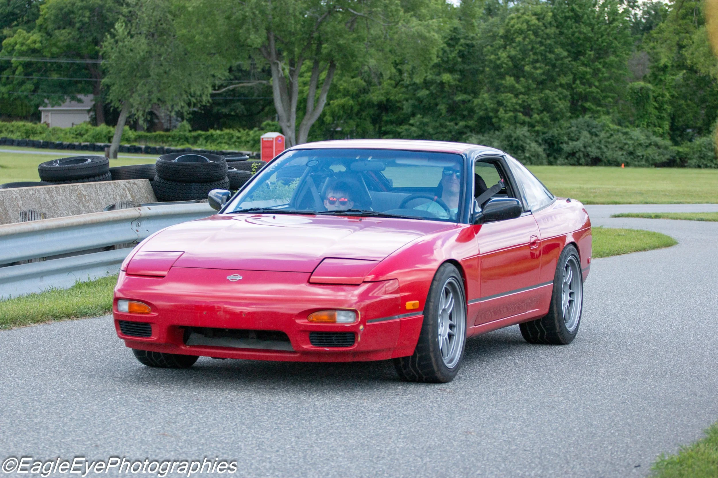 Red sports car driving on a paved road with trees and stacks of tires in the background.