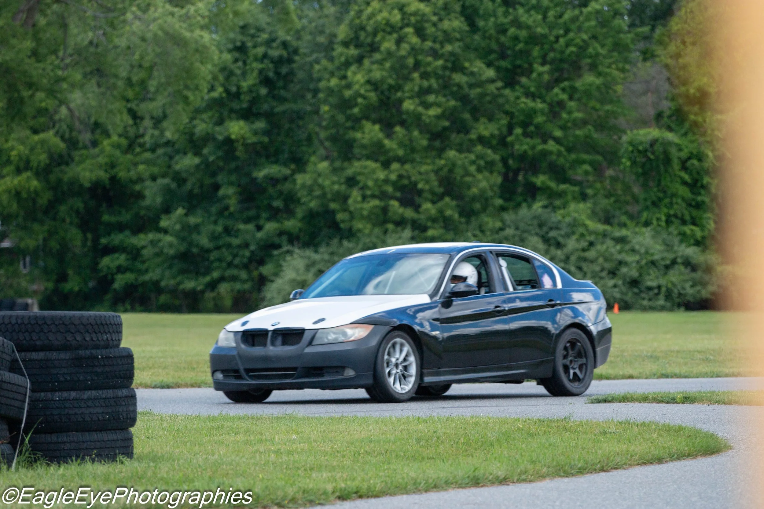 A blue and white race car on a track with green trees in the background, tire stacks on the left, and grass in the foreground.