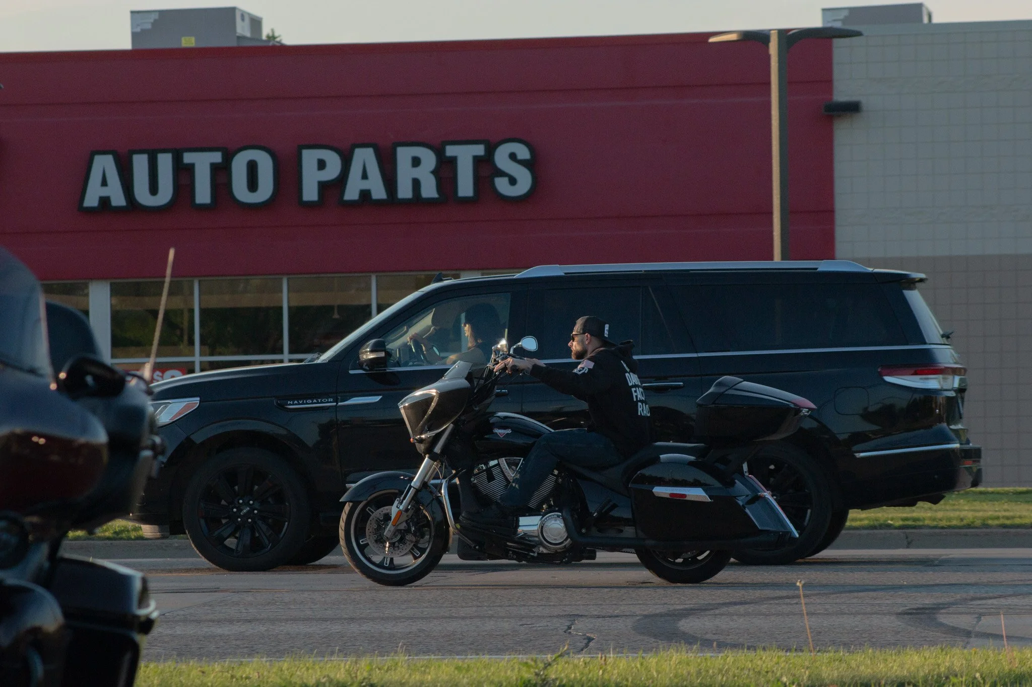 A store with large sign reading 'AUTO PARTS' in the background, a black SUV driving past, and a person riding a motorcycle in the foreground, in a parking lot during daytime.