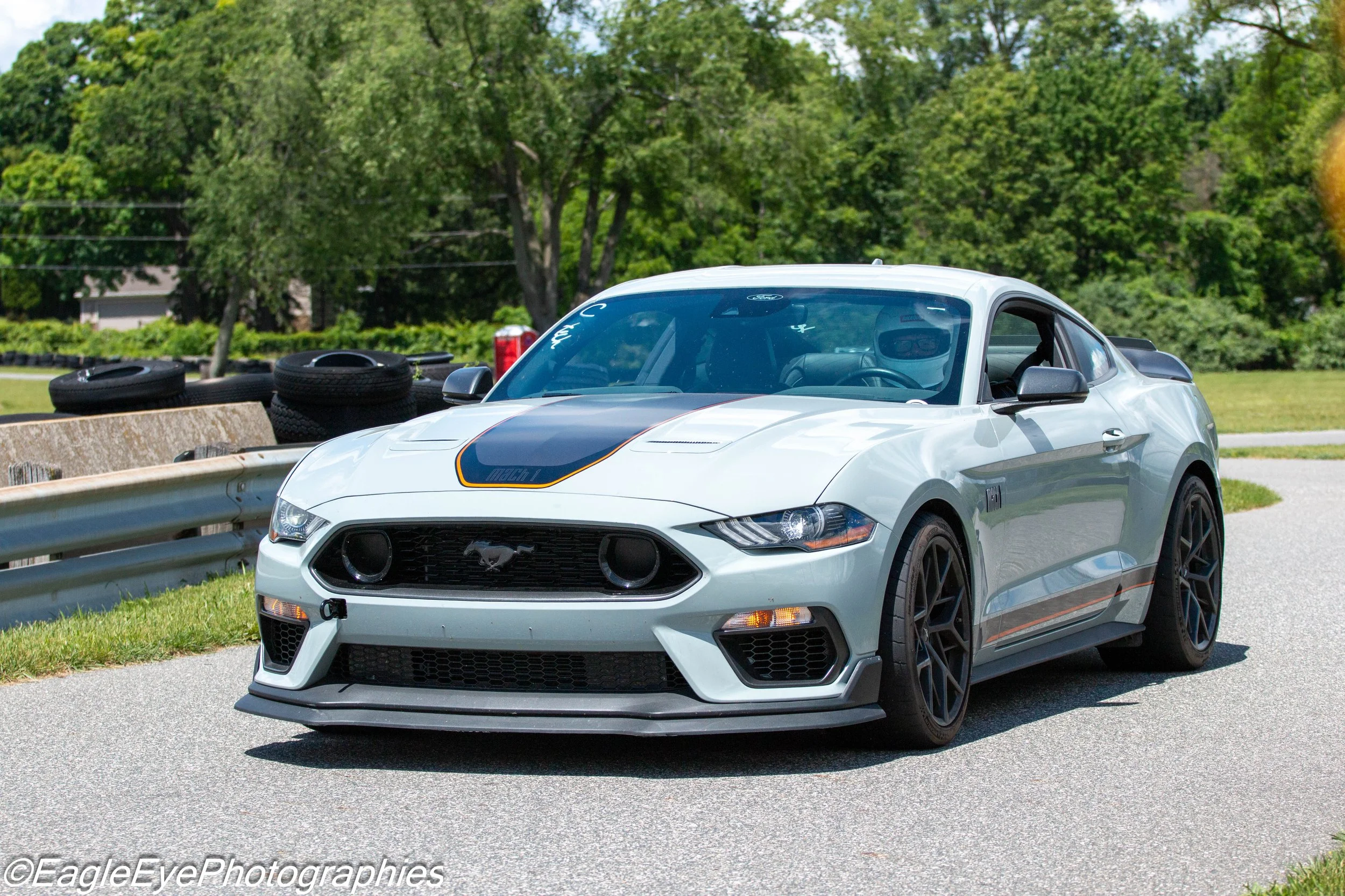A white Ford Mustang sports car with black racing stripes parked on the side of a road. There are trees in the background and tires stacked near a concrete barrier.