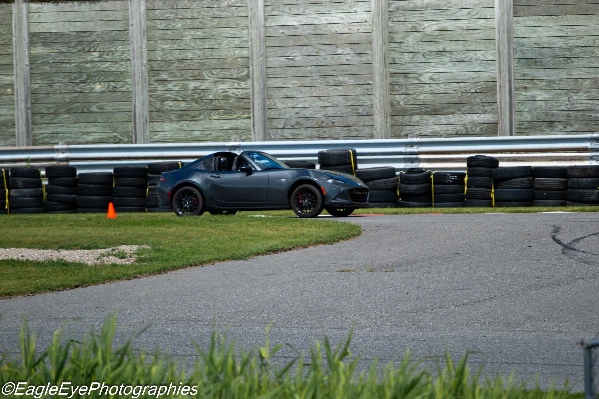 A gray sports car parked behind a tire barrier on a race track with grass and pavement, and a wooden fence in the background.
