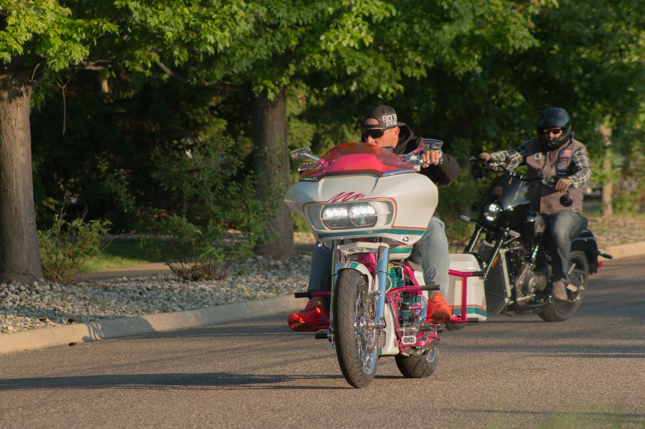 Two men riding motorcycles on a street with trees in the background. One rider is on a pink and white tricycle motorcycle, wearing sunglasses and a black hat with text, while the other rider is on a black motorcycle, wearing sunglasses and a helmet.