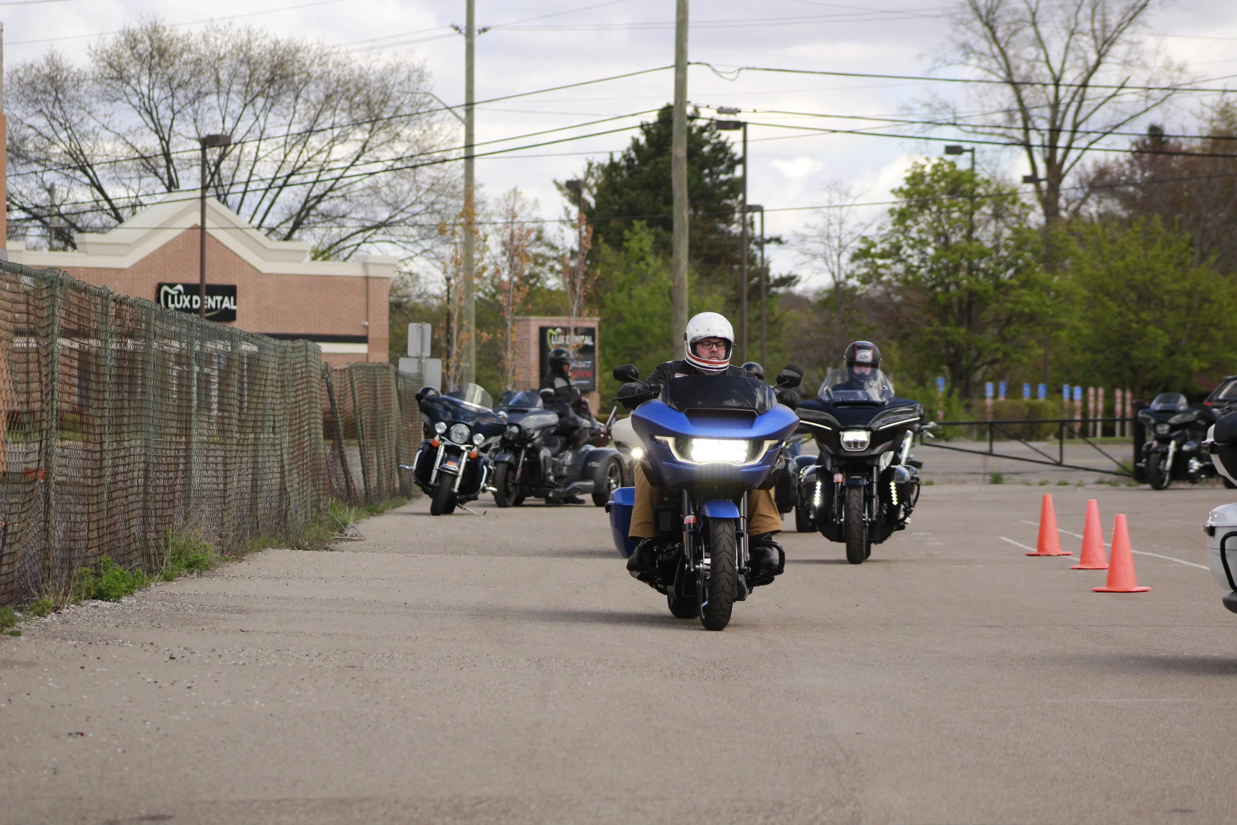 Group of motorcyclists riding together in a parking lot with orange traffic cones, trees, and a building in the background.