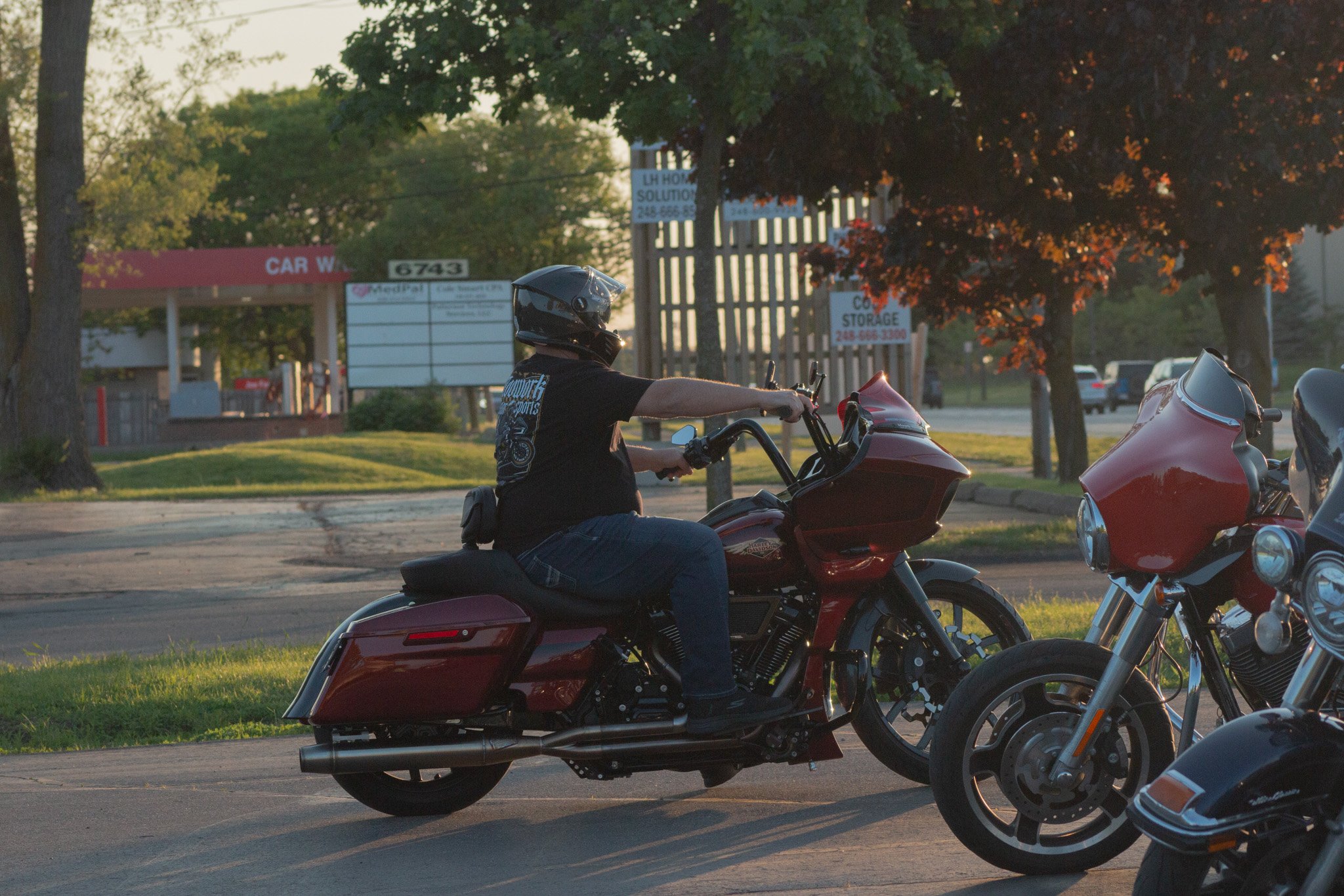 A person wearing a helmet and black T-shirt riding a red motorcycle with black accents on a street during sunset, with several other motorcycles parked nearby and trees in the background.