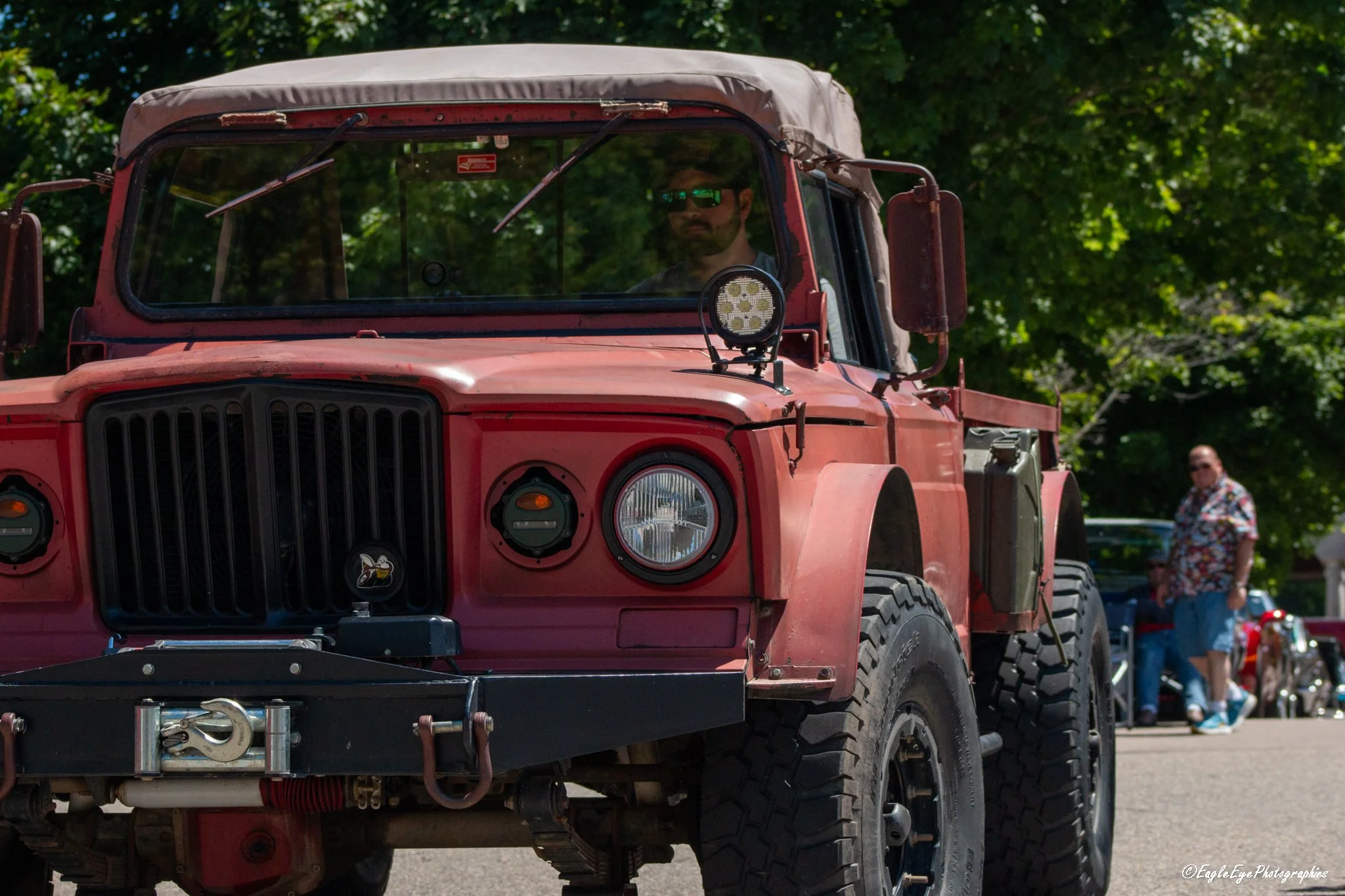 A person with sunglasses and a beard sitting inside a vintage red Jeep with a soft top, parked outdoors among green trees. In the background, a man wearing a colorful shirt and sunglasses stands near other vehicles and people.
