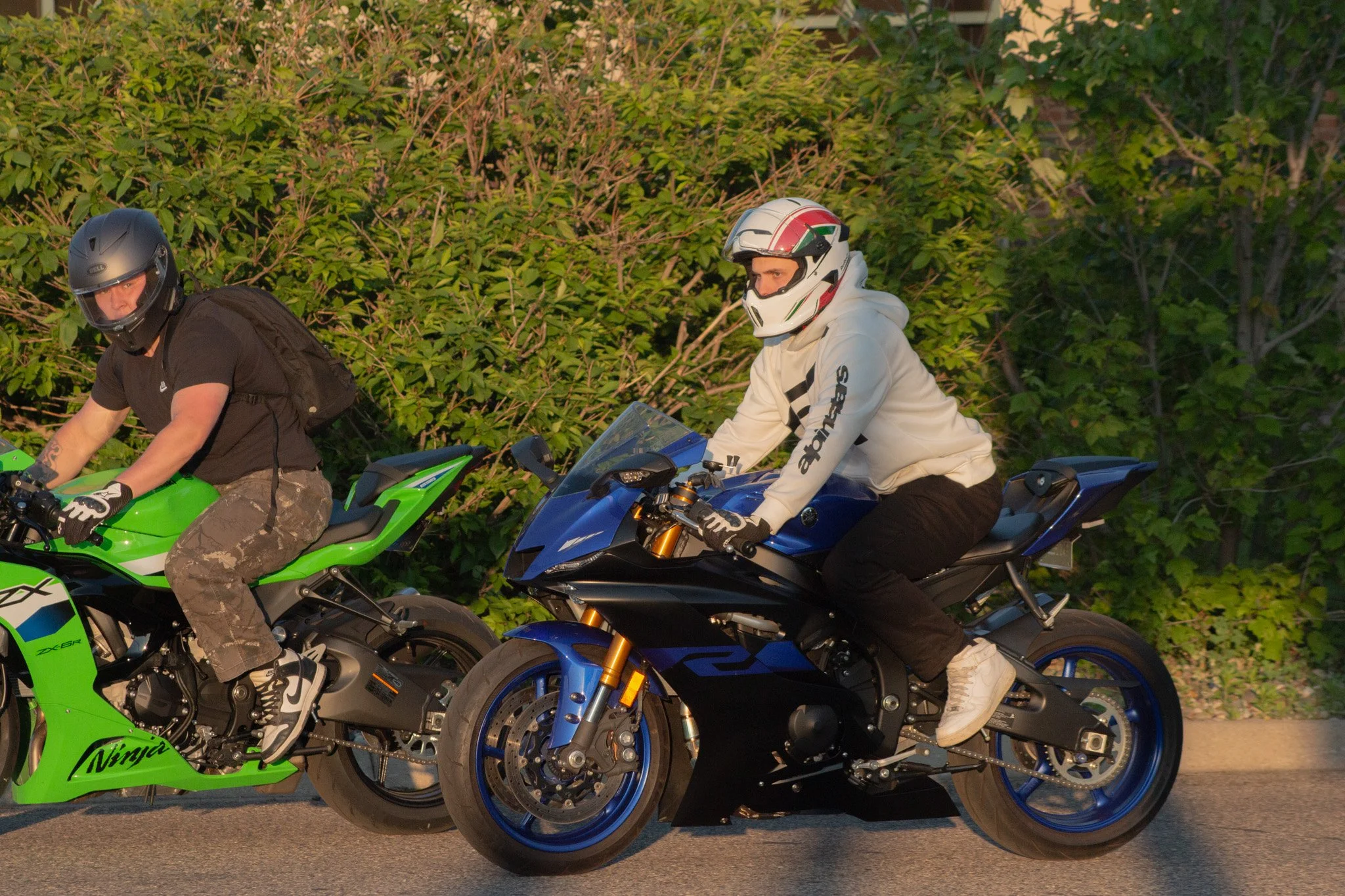 Two individuals riding motorcycles, one in black and the other in white, against a backdrop of green bushes. The rider in white wears a full-face helmet with red, green, and black accents, while the rider in black has a solid black helmet. The motorc