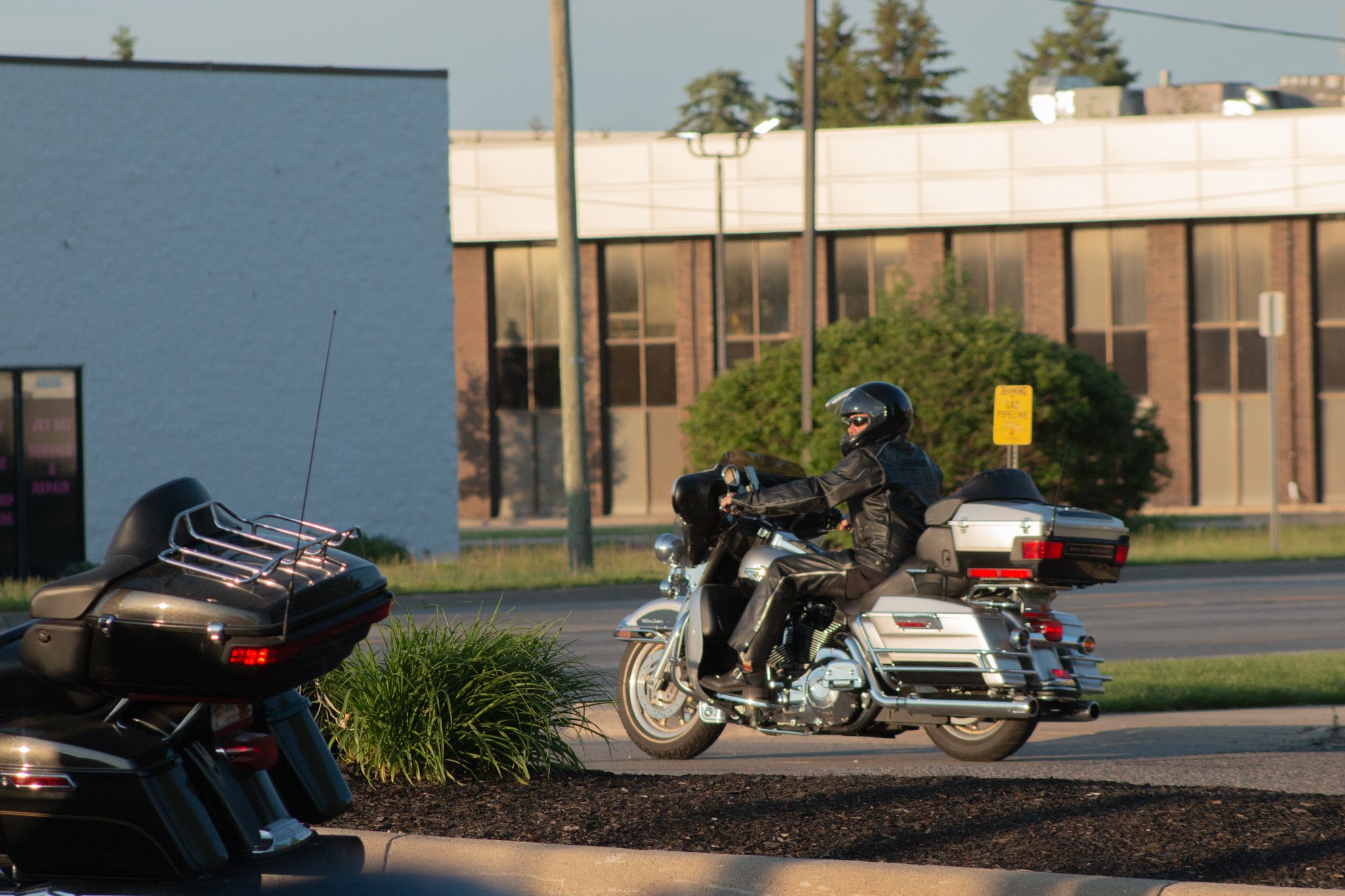 A motorcyclist wearing a helmet and black riding gear riding on a paved street, with a building, trees, and a yellow caution sign in the background.