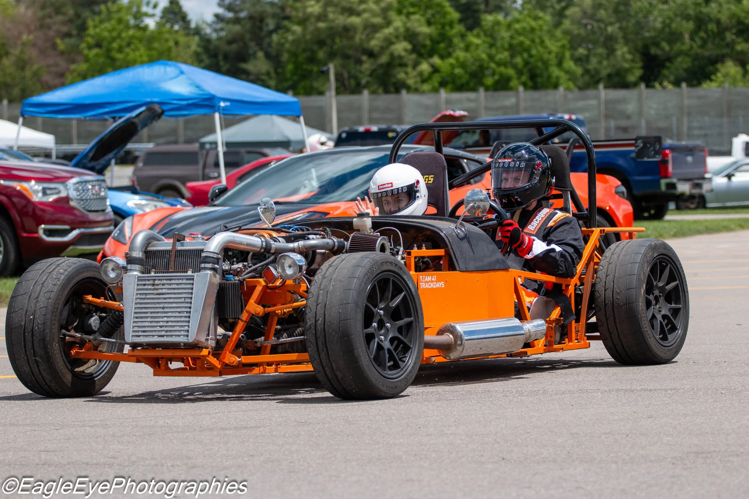 A go-kart with two racers wearing helmets, parked in a lot with cars and tents in the background.