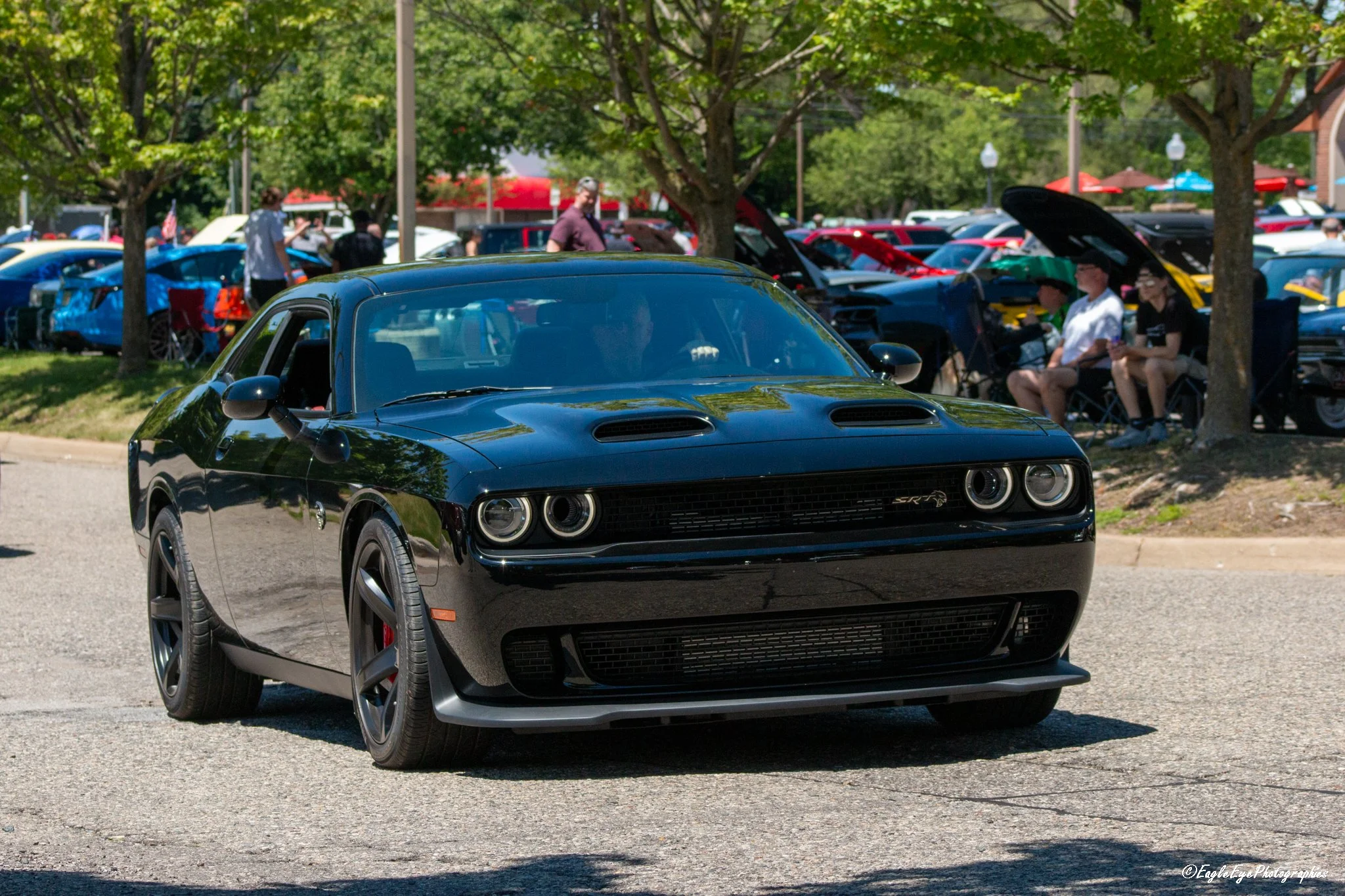 A black Dodge Challenger muscle car parked at a car show with other cars and people in the background under trees.