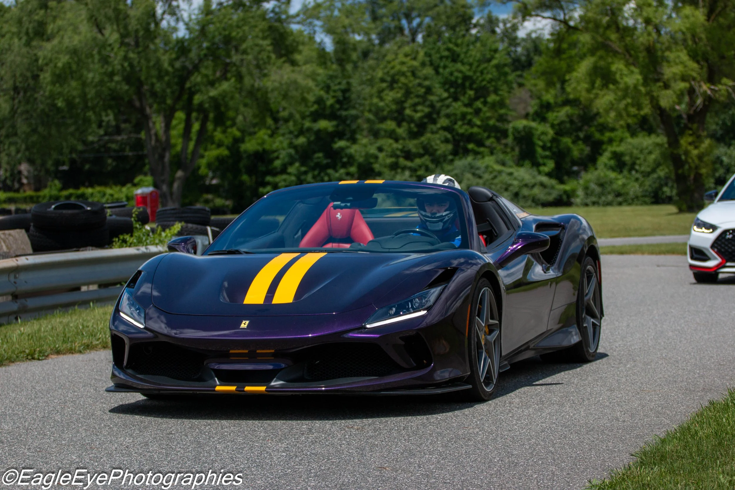 A black sports car with yellow racing stripes, a Ferrari logo, and a red racing seat inside, driven by a person wearing a helmet, on a road with green trees in the background.