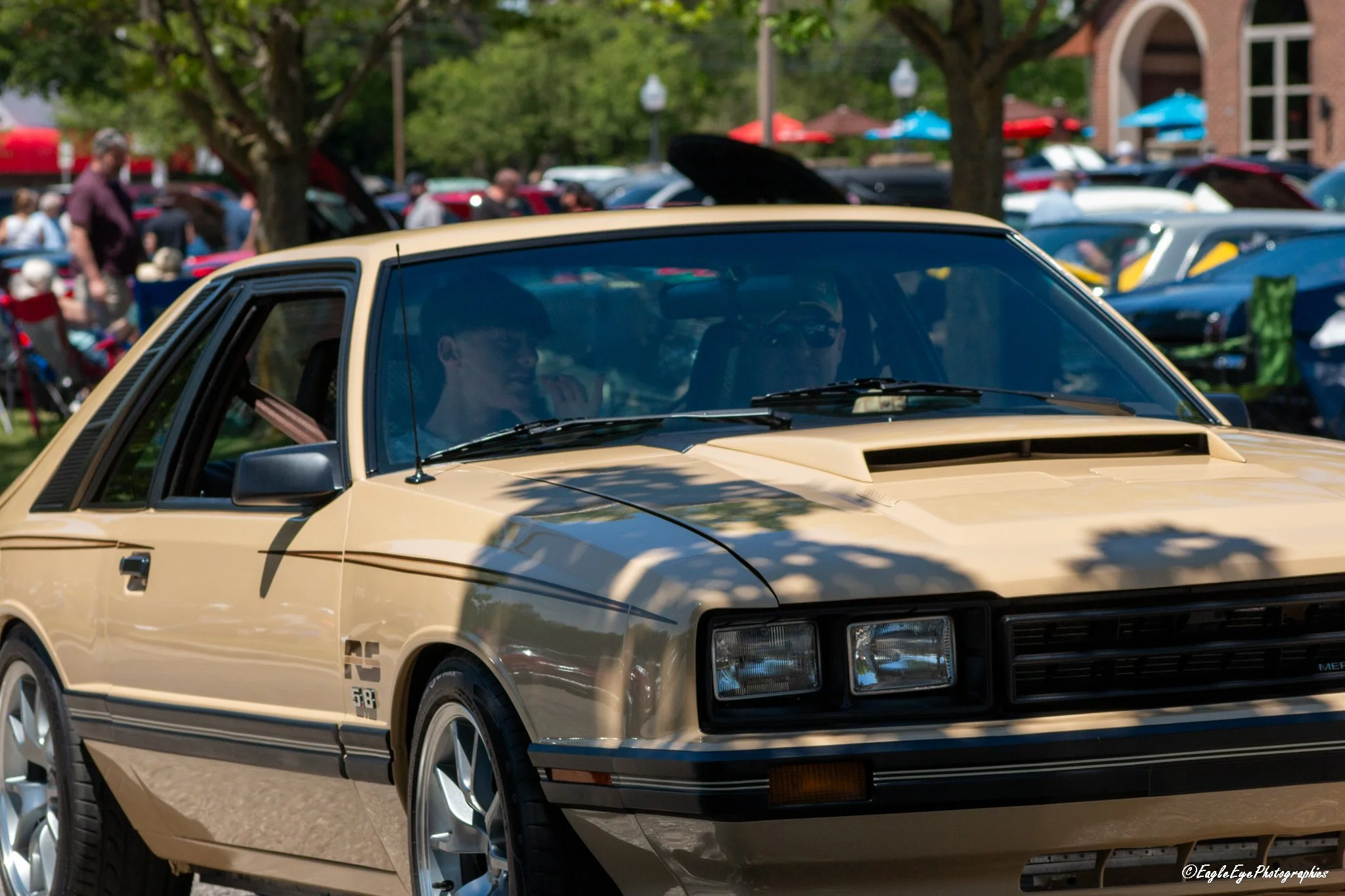 A beige vintage sports car parked at a car show with other vehicles and people in the background.
