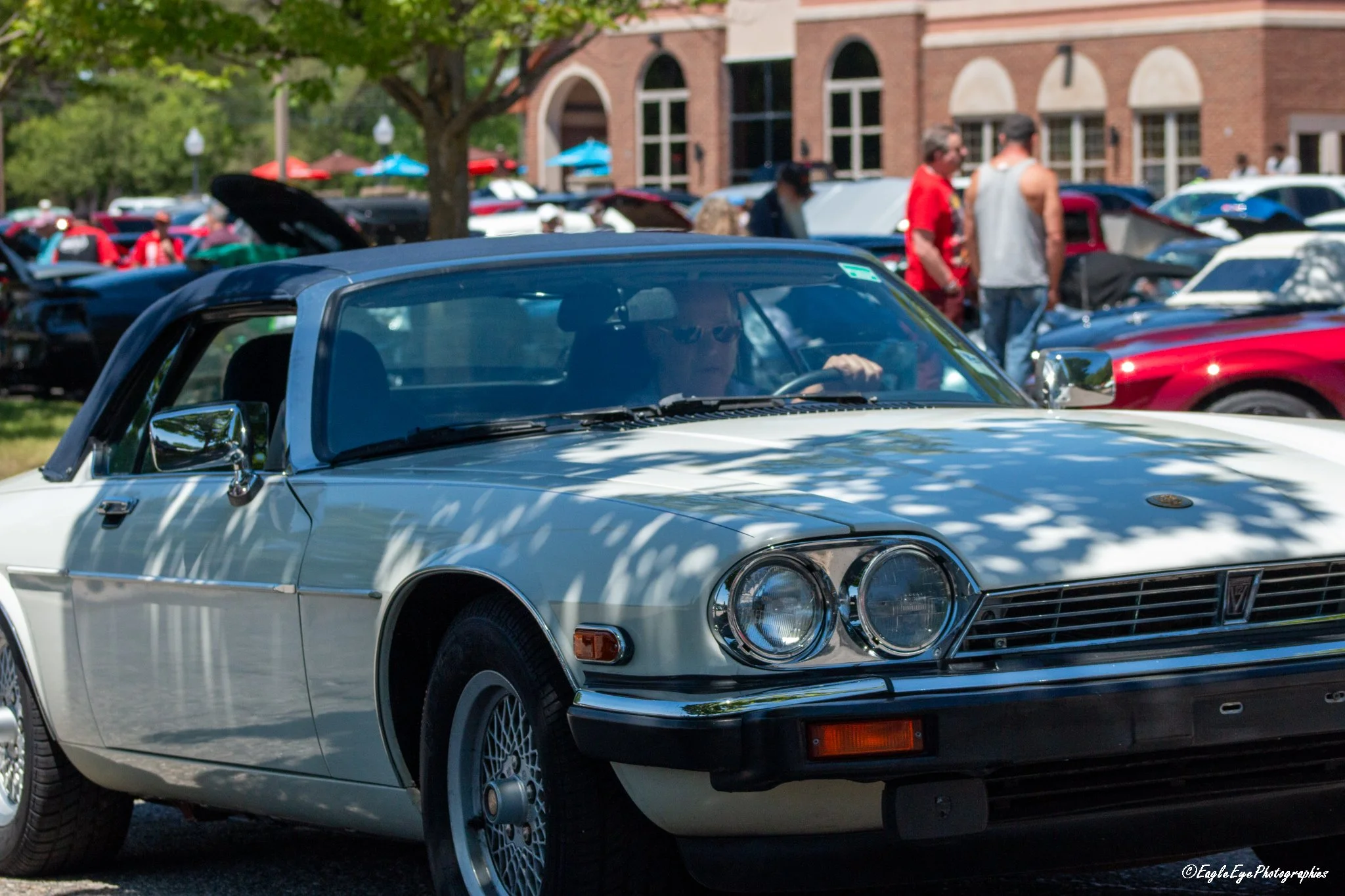 A white vintage sports car with a blue roof parked in a lot filled with other classic and modern cars at a car show.