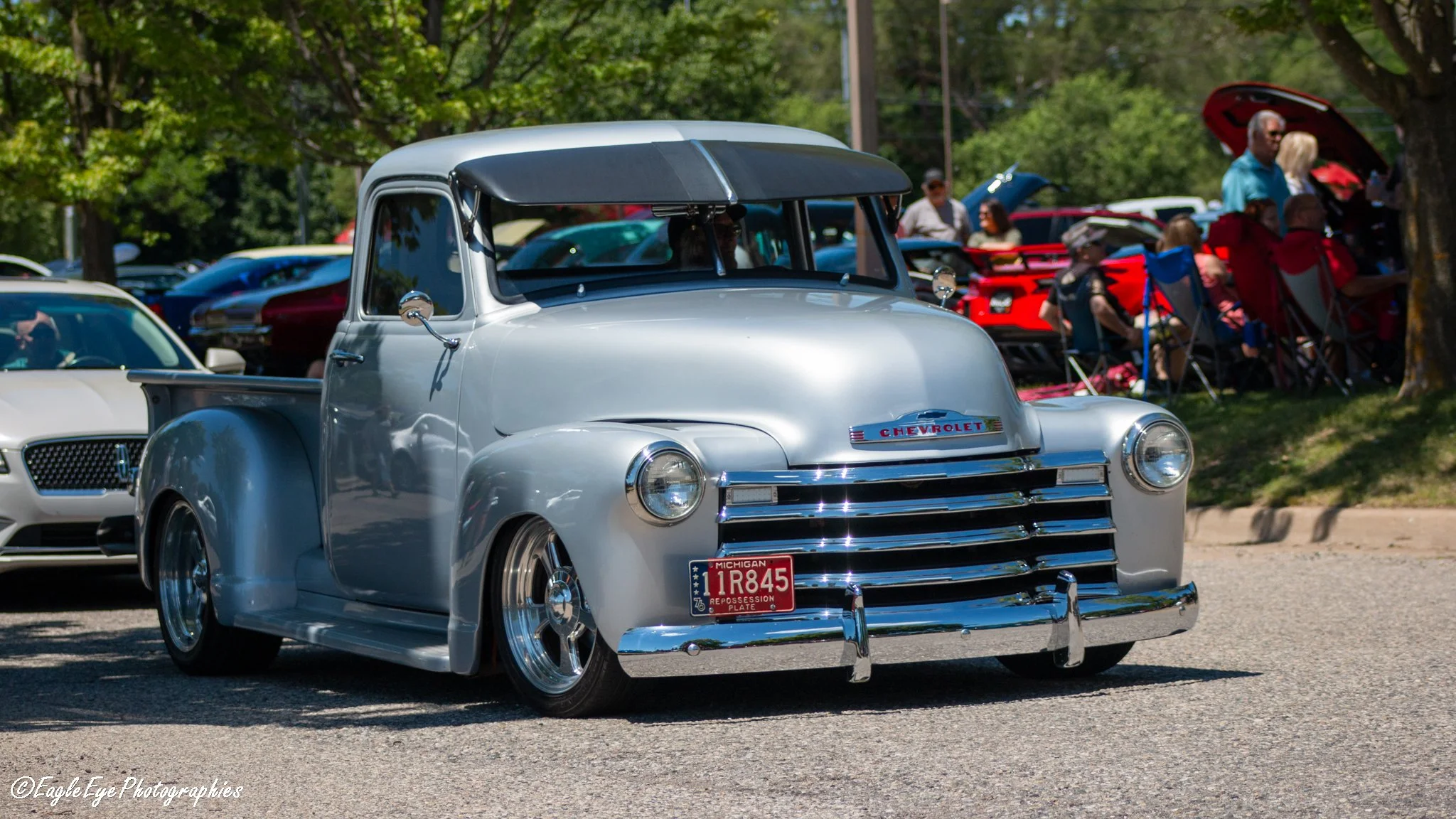 A vintage Chevrolet pickup truck with a silver body and black roof parked at a car show. The truck has a Michigan license plate reading 1R845 and is surrounded by other classic cars and onlookers.