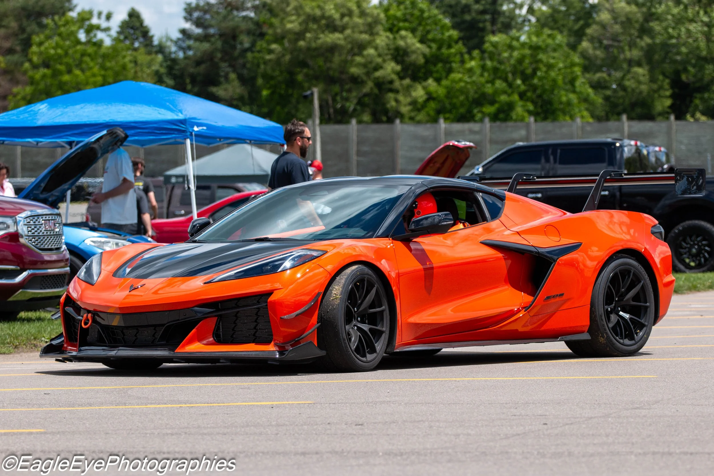Bright orange sports car with black accents parked at an outdoor car show. People walk around, some inspecting cars under a blue canopy, with green trees in the background.