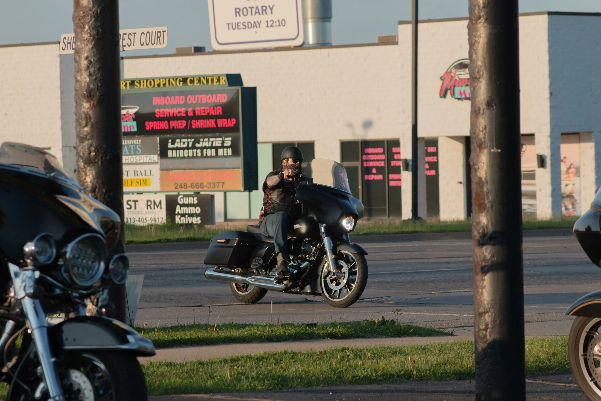 A person riding a black motorcycle on the road, wearing a helmet and sunglasses, with storefronts and signs in the background.