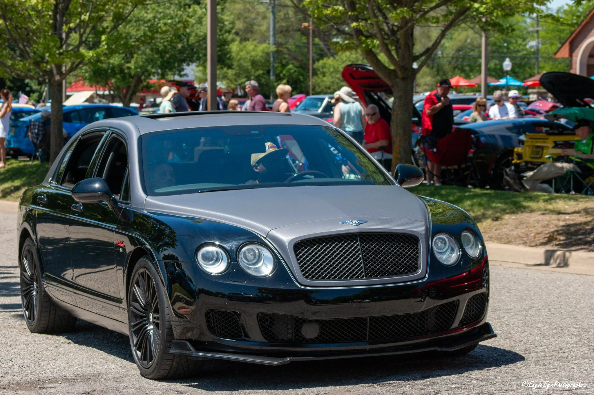 A black luxury Bentley car parked outdoors at a car show with people walking around and looking at other cars in the background.