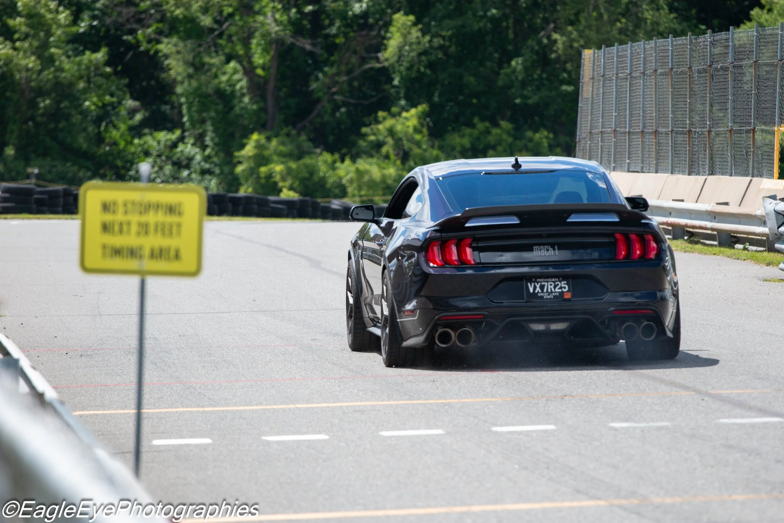A black Ford Mustang Mach 1 sports car driving on a racetrack with a yellow warning sign in the foreground and a chain-link fence on the right side, with green trees in the background.