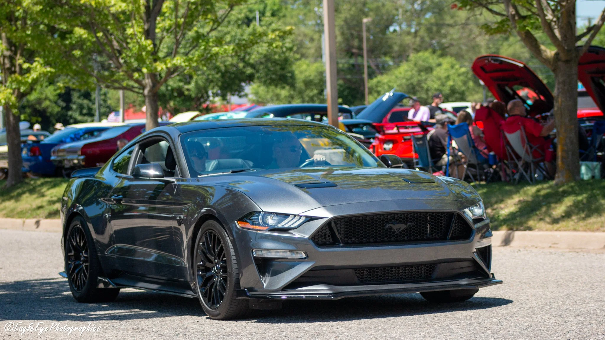 A dark gray Ford Mustang sports car driving past a group of people sitting on lawn chairs under trees at a car show.