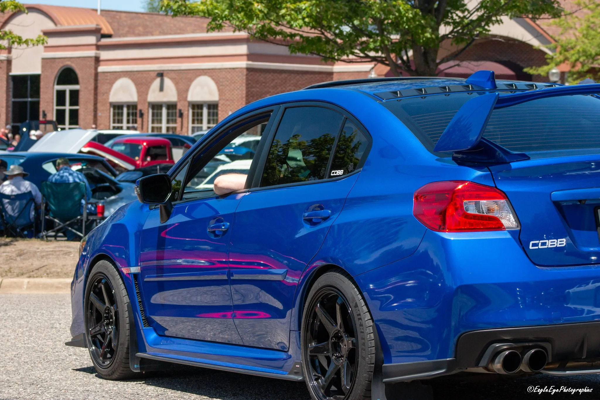 A blue sports car parked at an outdoor event with people and other vehicles in the background.