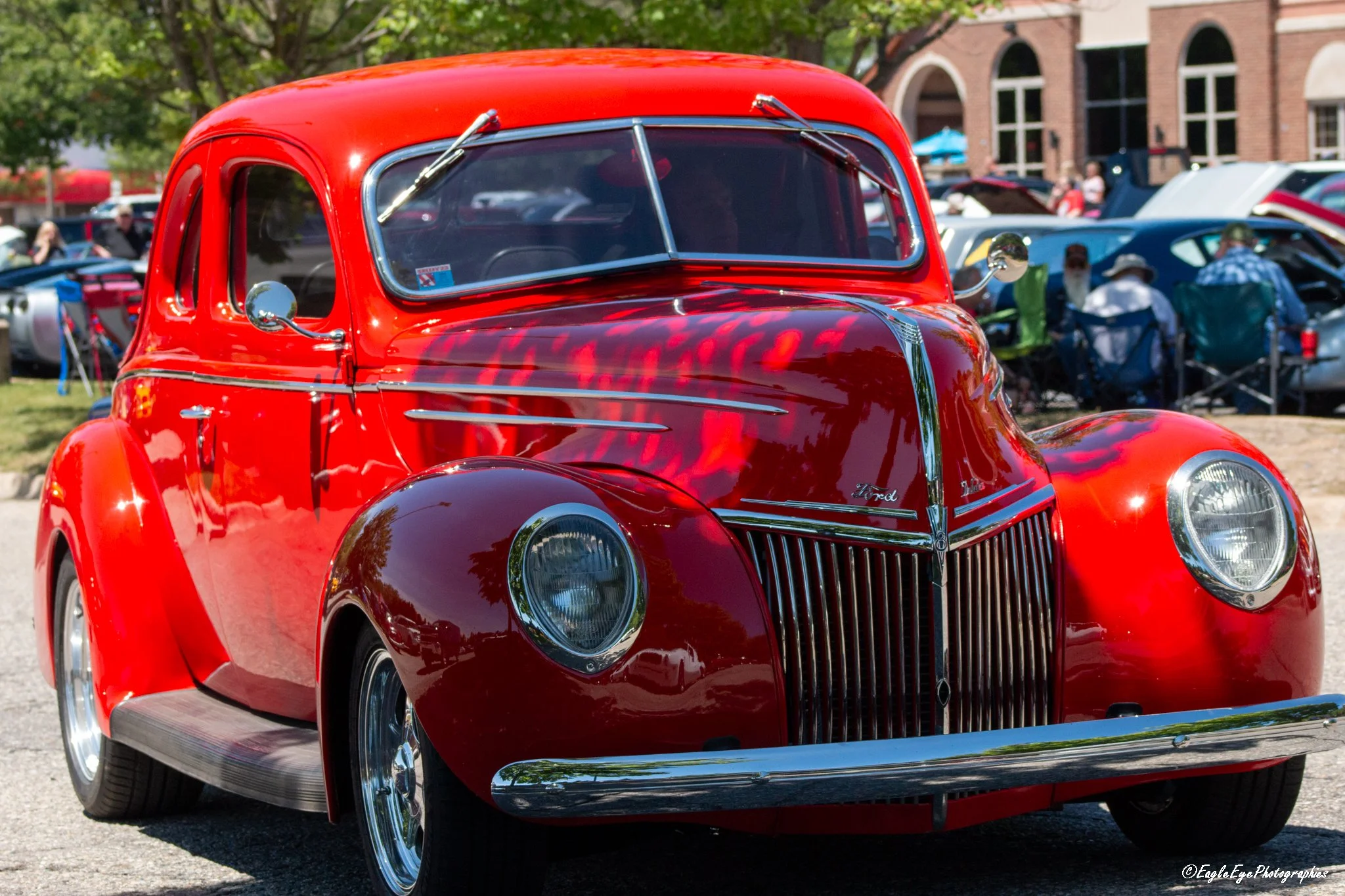 Close-up of a vintage red Ford car at a car show with other cars and people in the background.