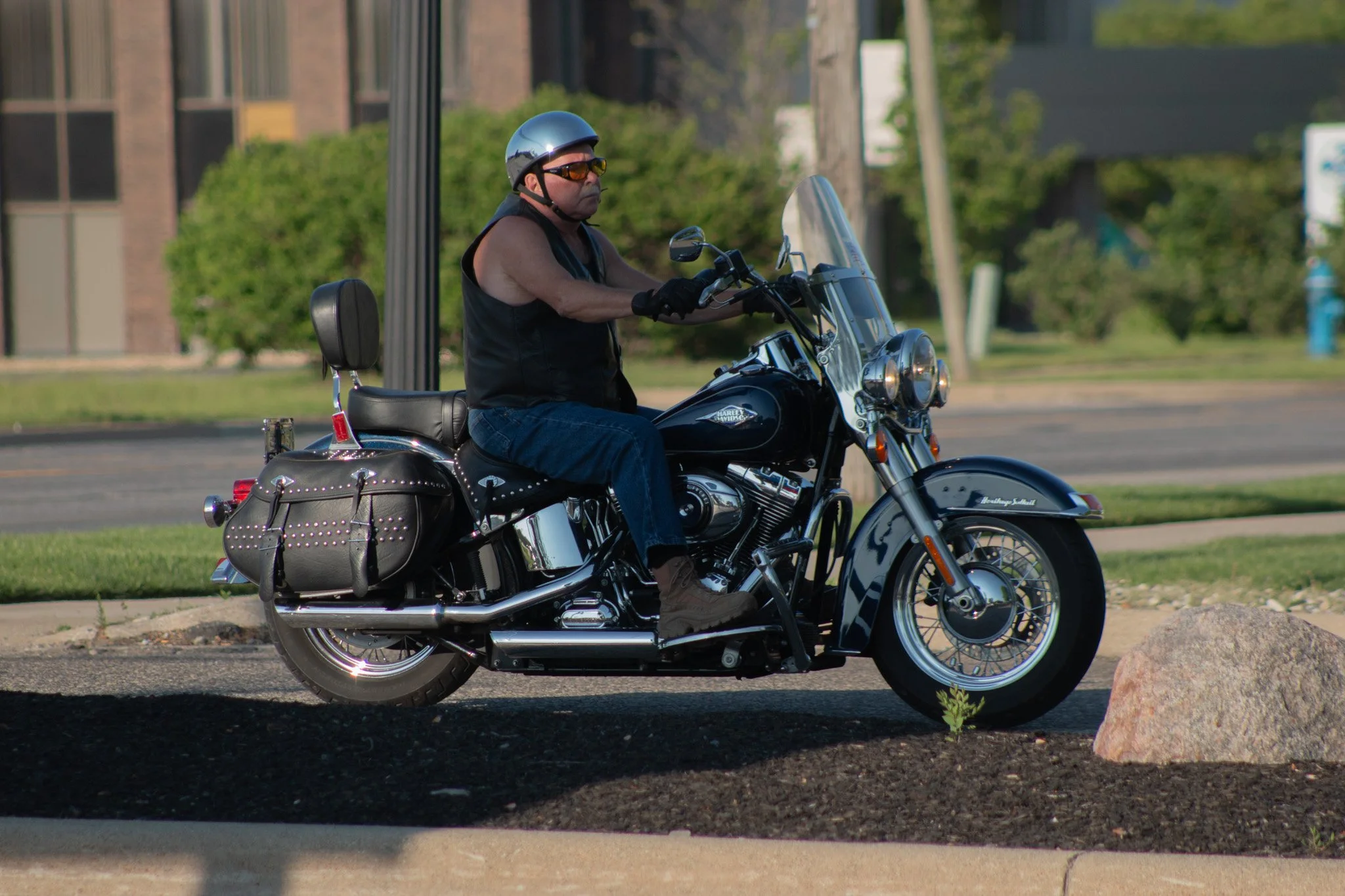A man riding a black Harley Davidson motorcycle on a city street, wearing a black helmet, sunglasses, a sleeveless black vest, jeans, gloves, and brown boots.