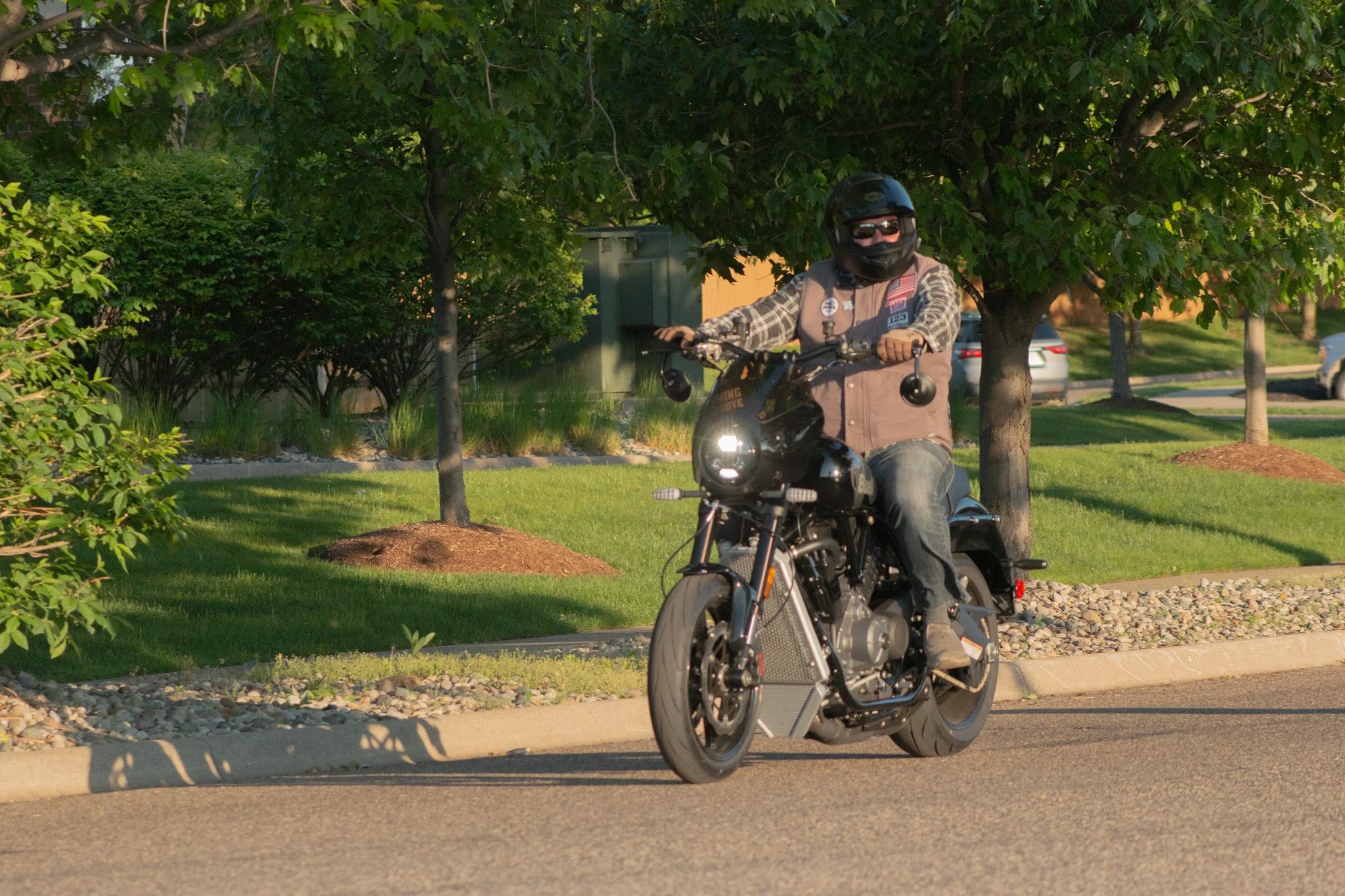 A person riding a black motorcycle on a city street, wearing a black helmet, sunglasses, a beige vest, and plaid shirt, with green trees and parked cars in the background.