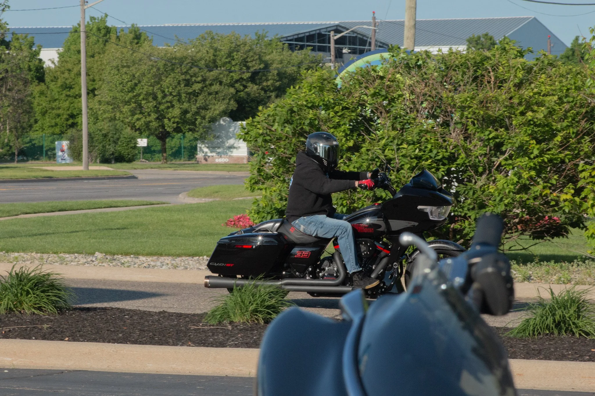 Person riding a black motorcycle with a helmet, black jacket, and jeans on a sidewalk with greenery and trees, in an urban area with industrial buildings and power lines in the background.
