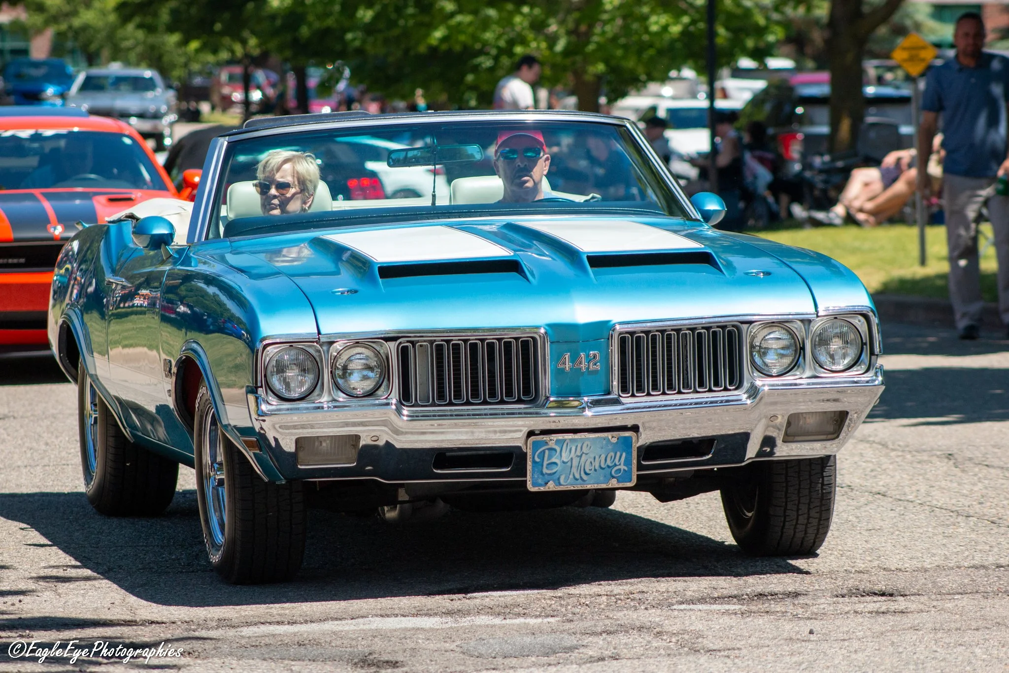 A blue classic car with white racing stripes, a license plate reading 'Blue Money,' driven by a man and woman, at an outdoor car show with other cars and people in the background.