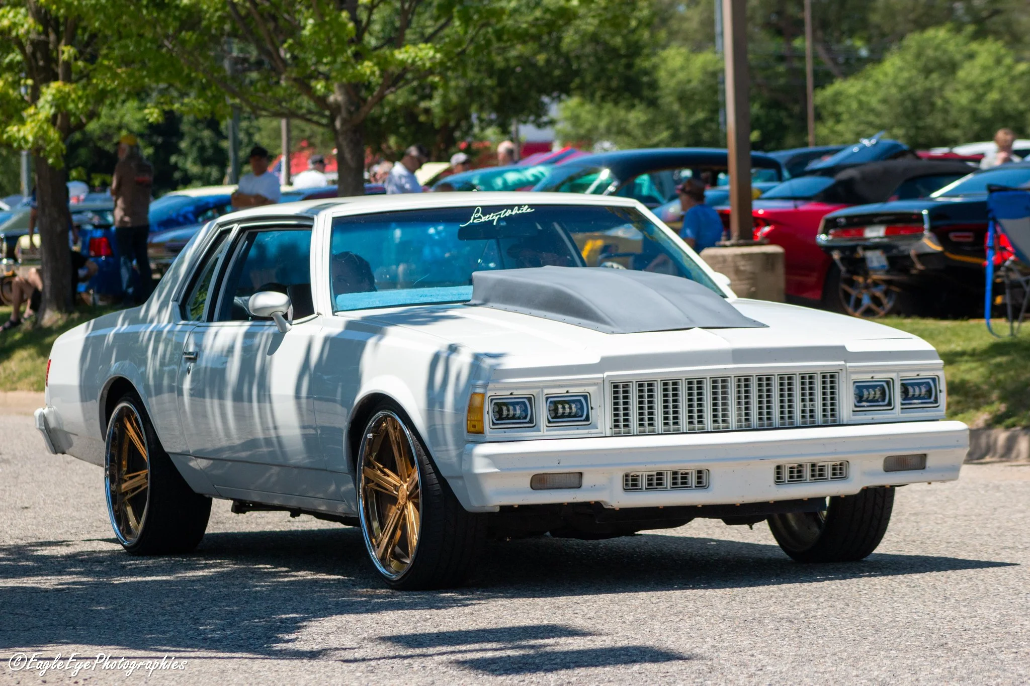 A white vintage car with gold rims participating in a car show, with a sign on the windshield that reads Betty White, parked outdoors with trees and a crowd in the background.