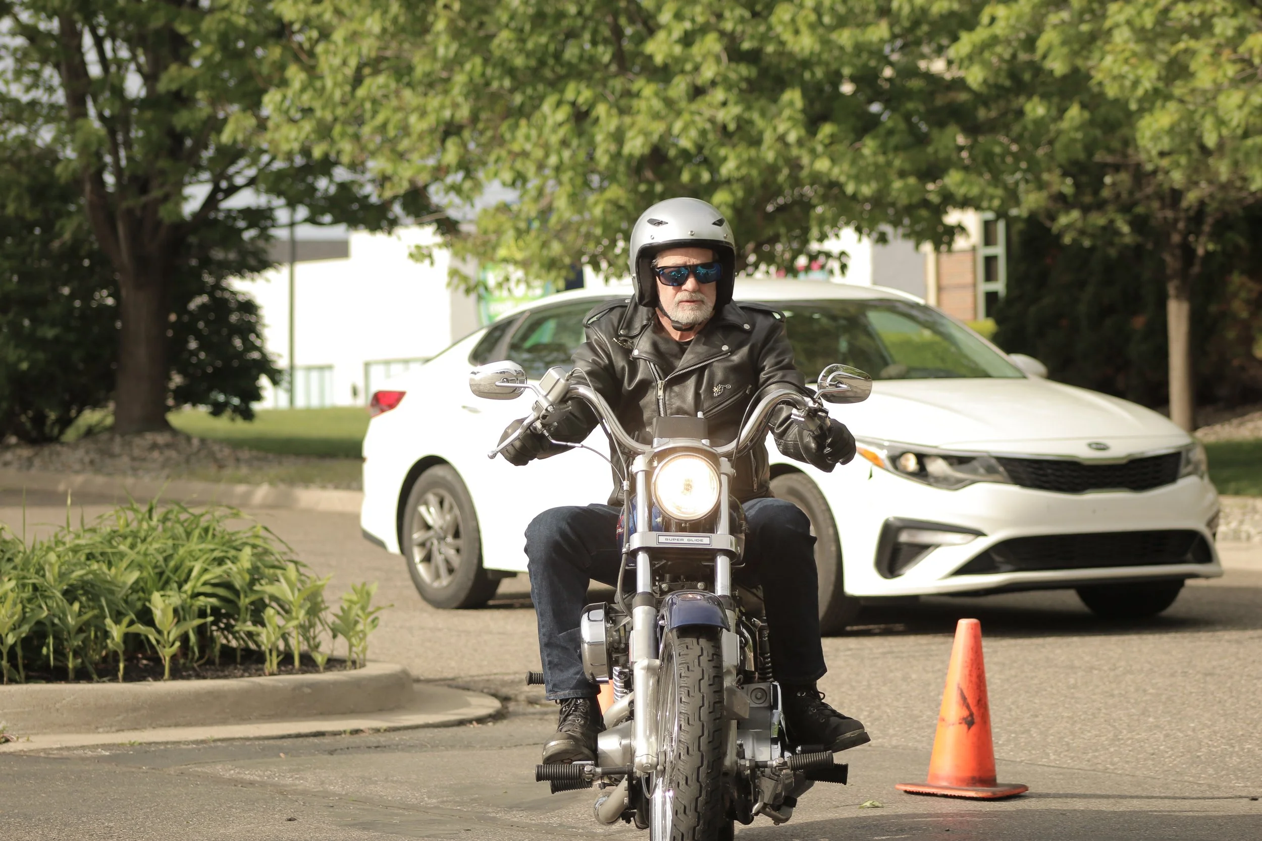 Older man wearing a helmet, sunglasses, and a leather jacket riding a motorcycle through a parking lot marked with orange cones. Green trees and a white car are visible in the background.
