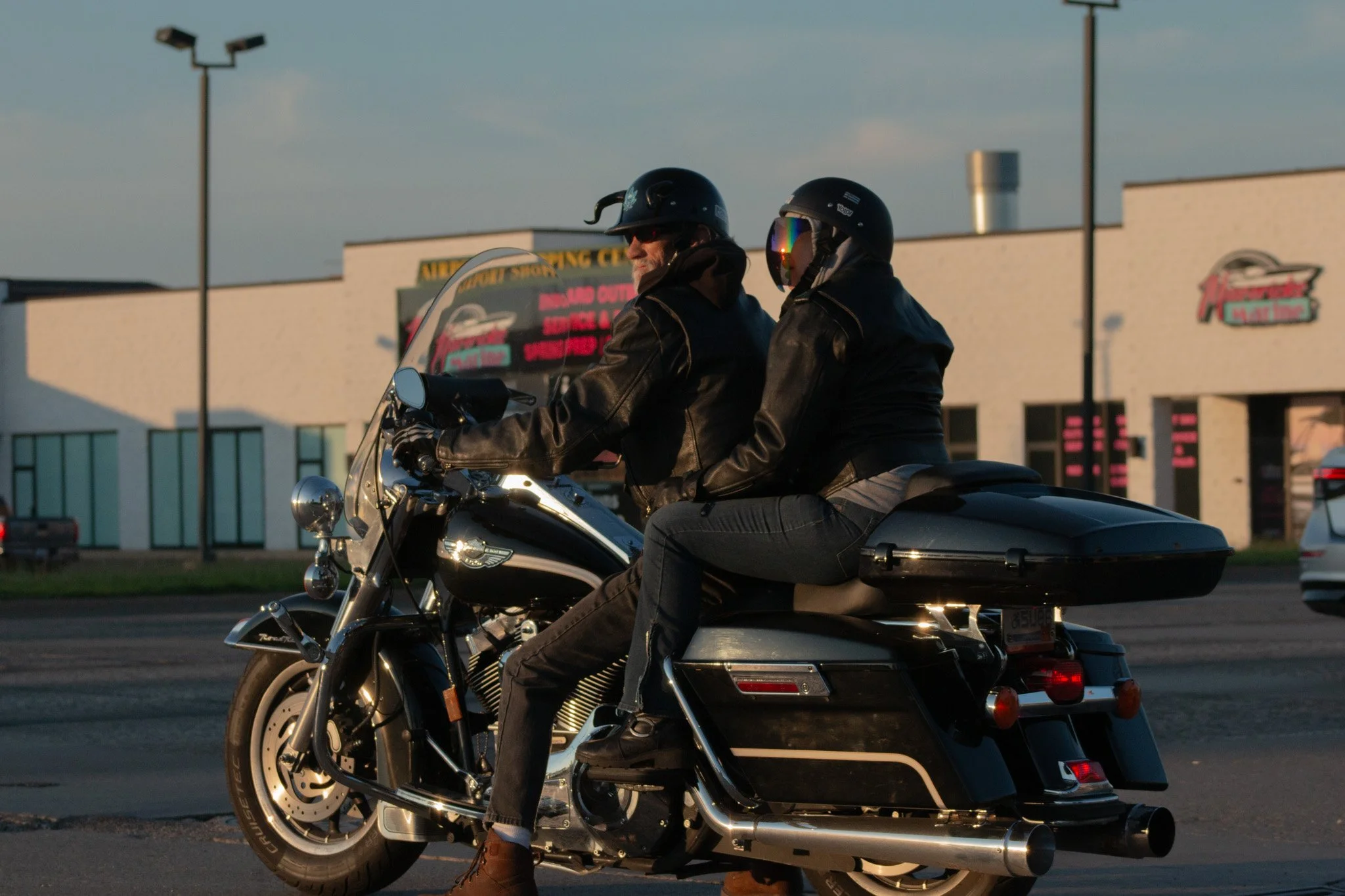 Two motorcyclists riding on a highway, both wearing black helmets and leather jackets, with a building and a billboard in the background during evening time.