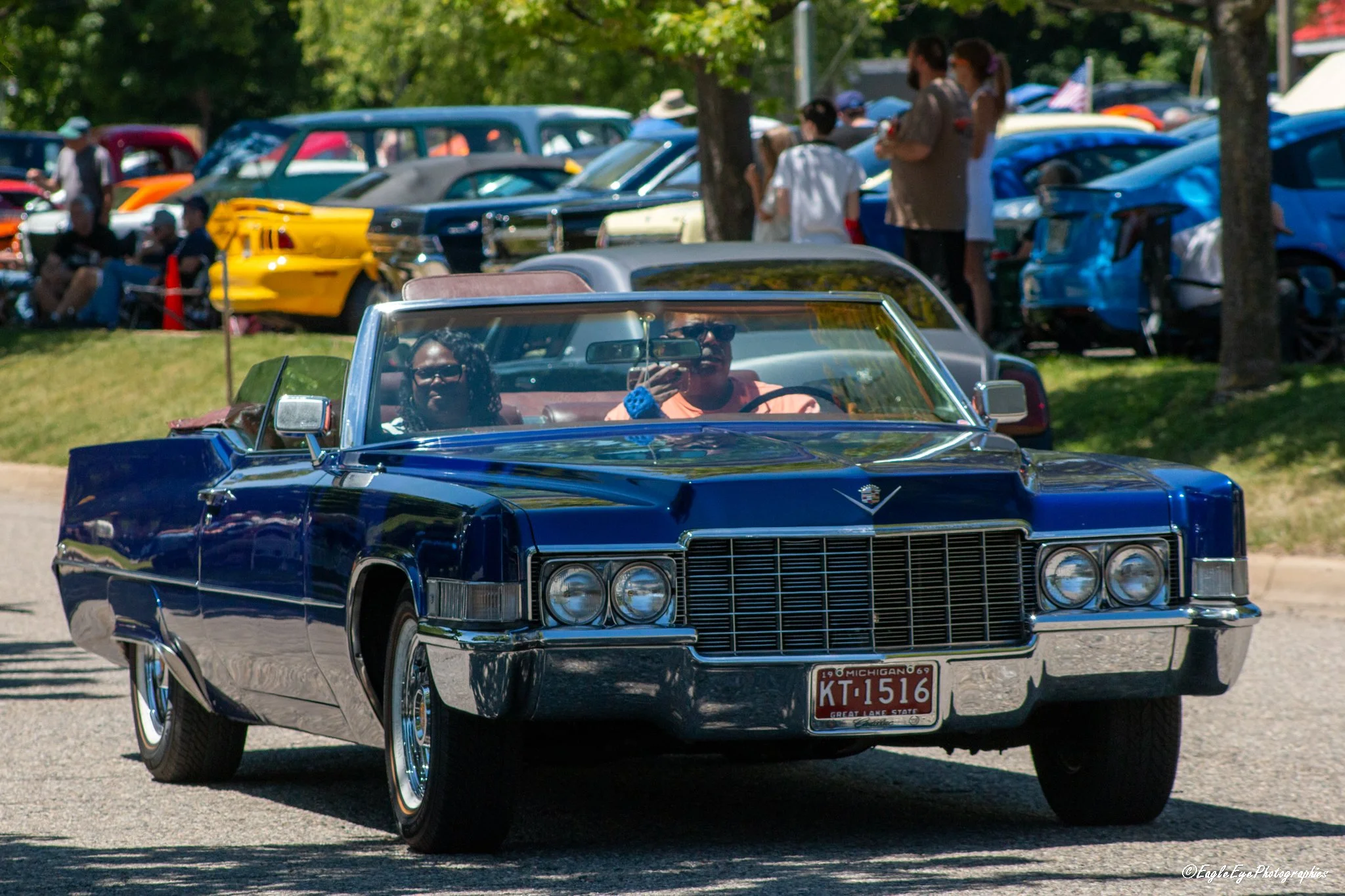 A vintage black Cadillac convertible car with a red license plate driving at a car show event. People and classic cars are visible in the background at an outdoor park setting.