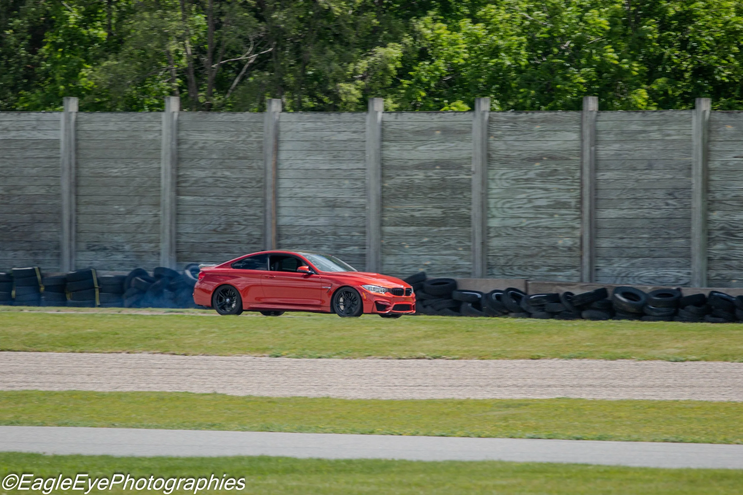 A red sports car driving on a racetrack with a tire barrier and a wooden fence in the background, and green trees beyond the fence.
