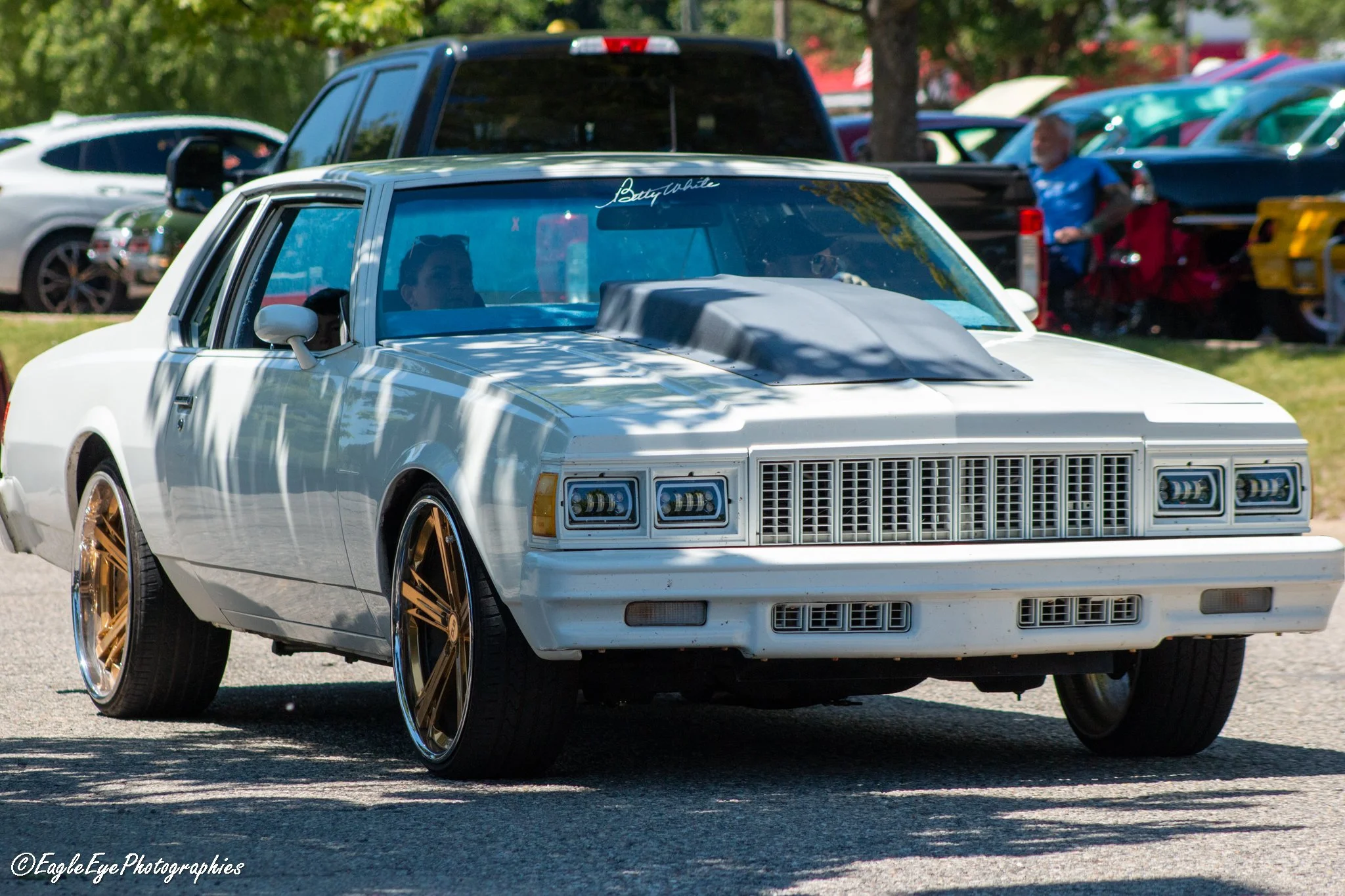 A white vintage car with gold rims parked outdoors, with people and other vehicles visible in the background at a car show.