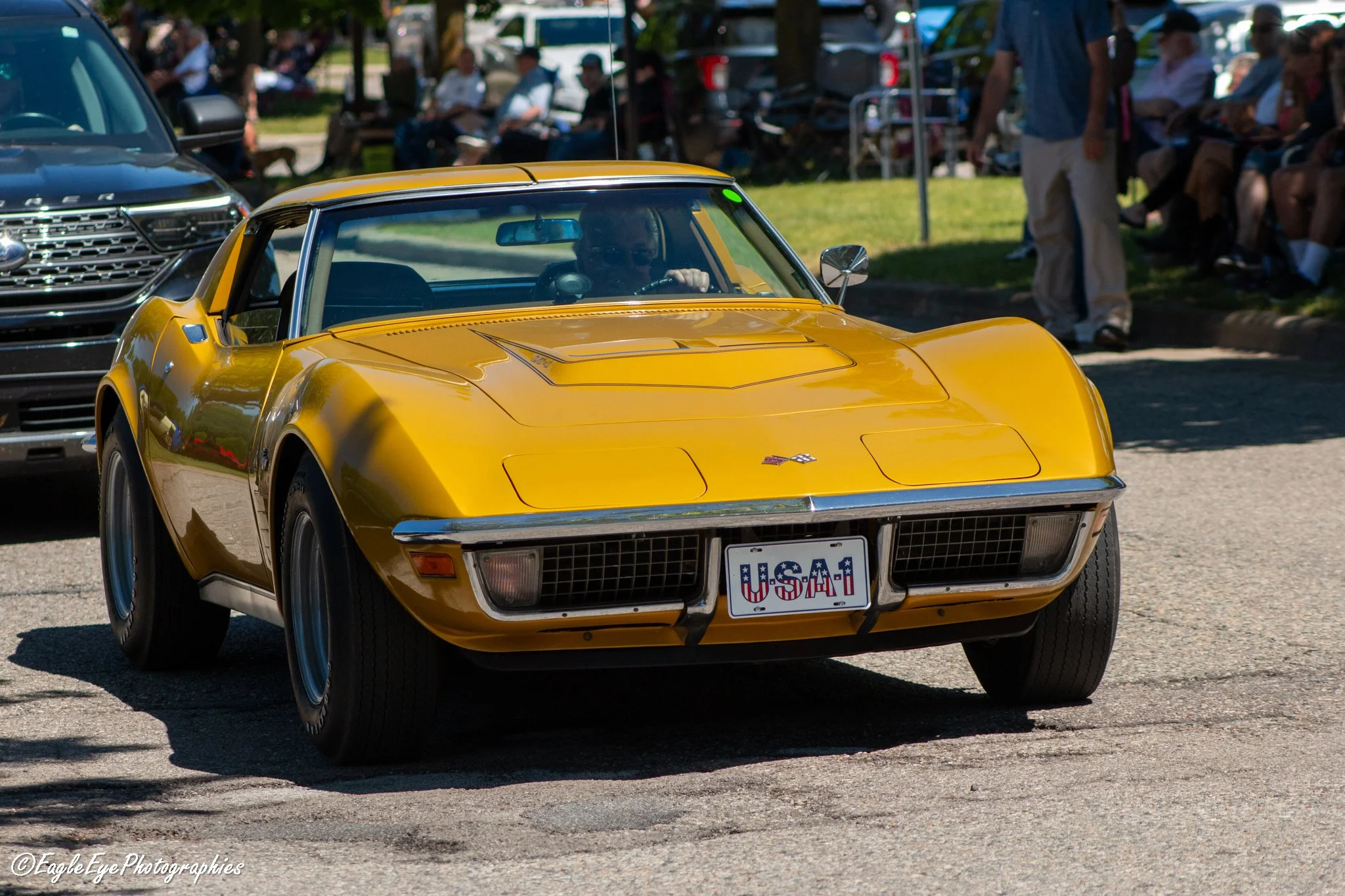 A yellow vintage Chevrolet Corvette with a USA license plate, driving on a street during a car show or parade. There are people sitting on benches and standing on the grass in the background, observing the event.