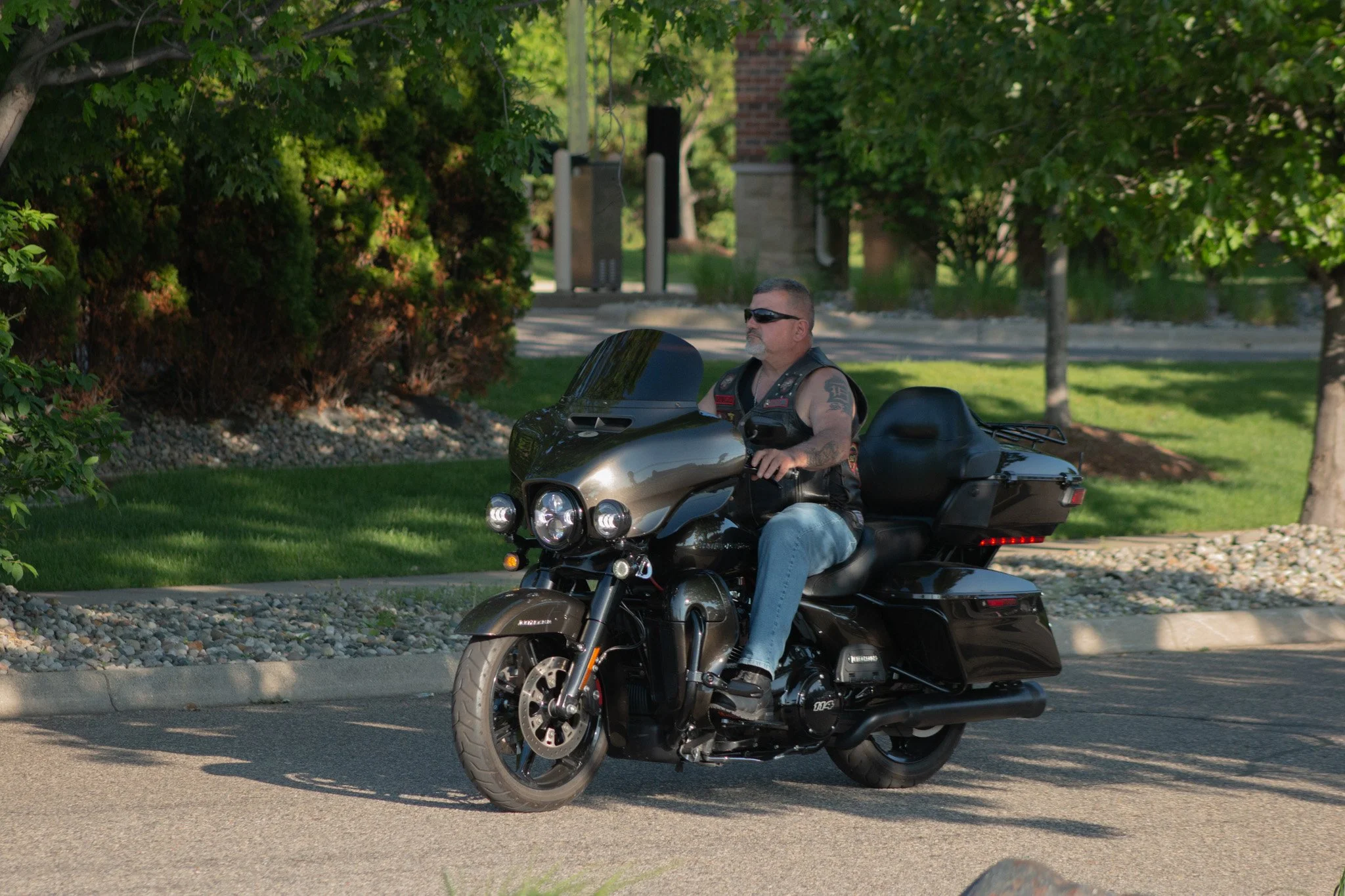 A man with tattoos, sunglasses, and a vest riding a black motorcycle with a large front fairing and luggage compartment, on a suburban street lined with trees and bushes.