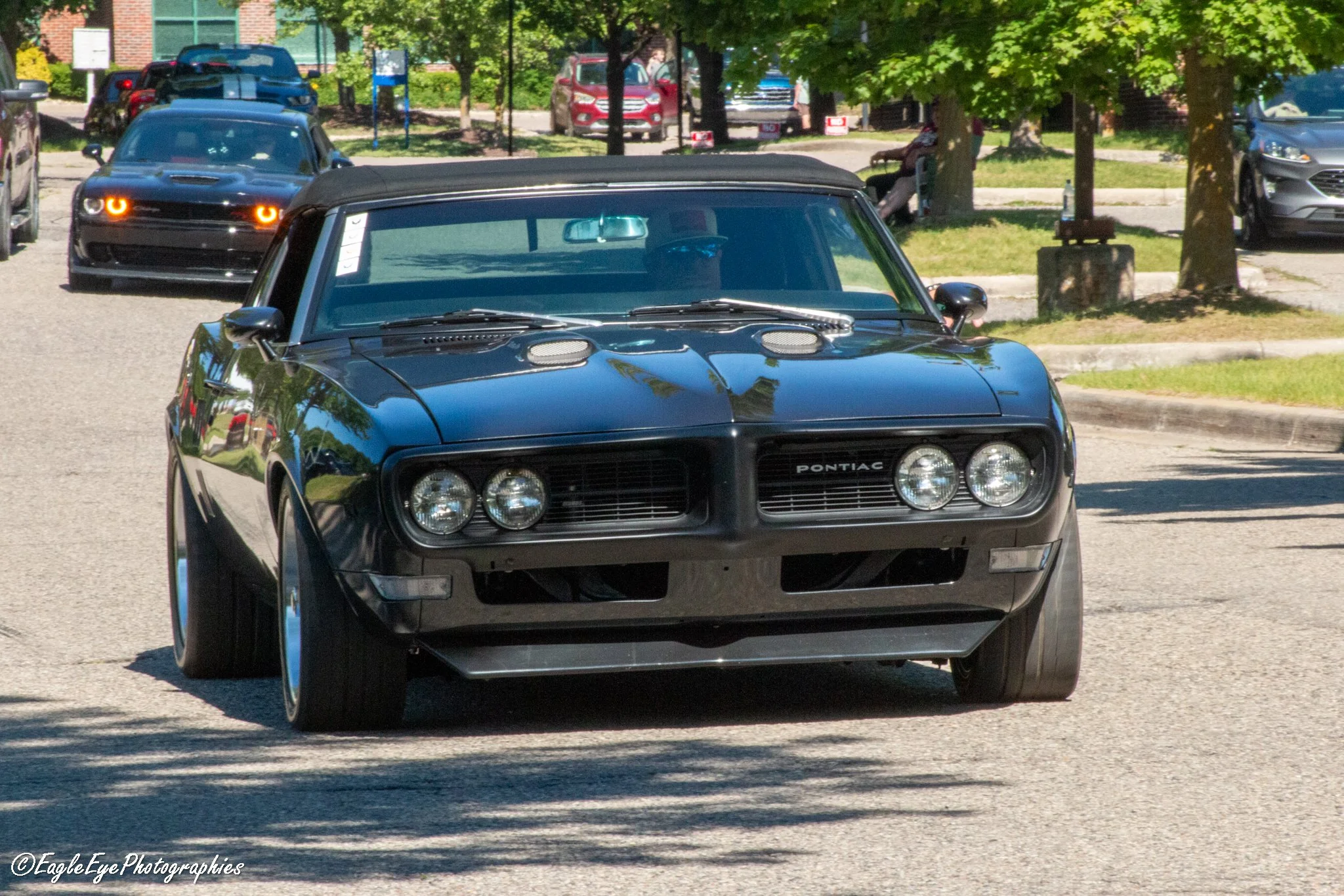 A black vintage Pontiac sports car driving on a street with bicycles, cars, and trees in the background during the daytime.